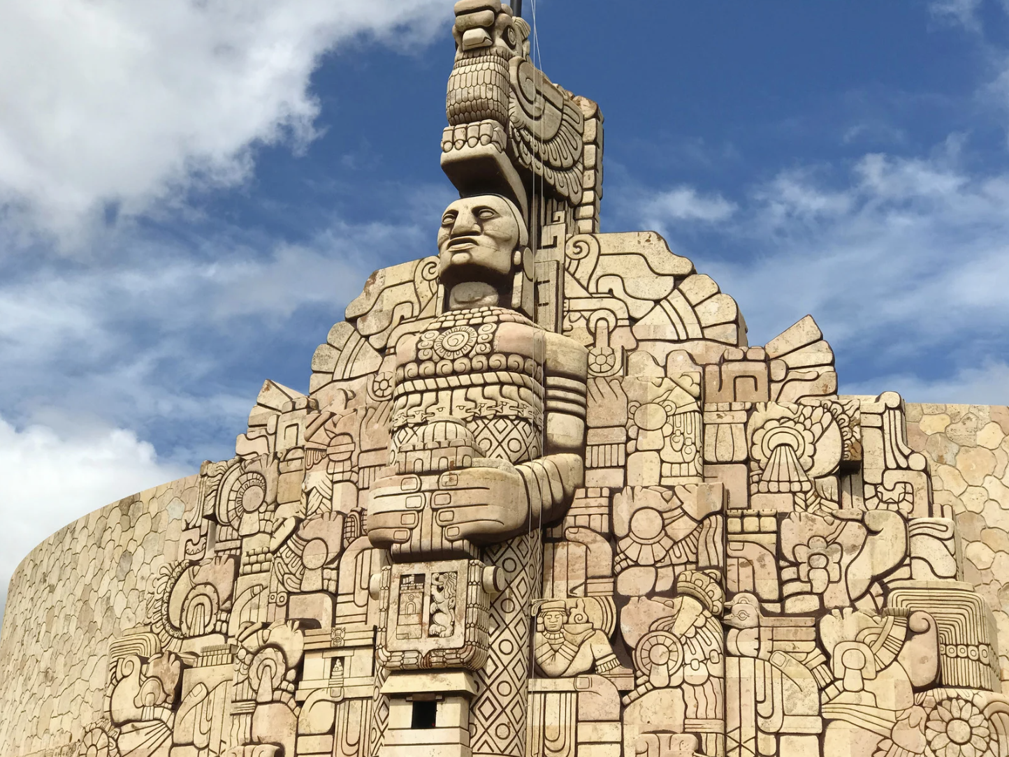 Escultura monumental de estilo prehispánico en color piedra, mostrando figuras humanas y atributos culturales, con un fondo de cielo azul con nubes.