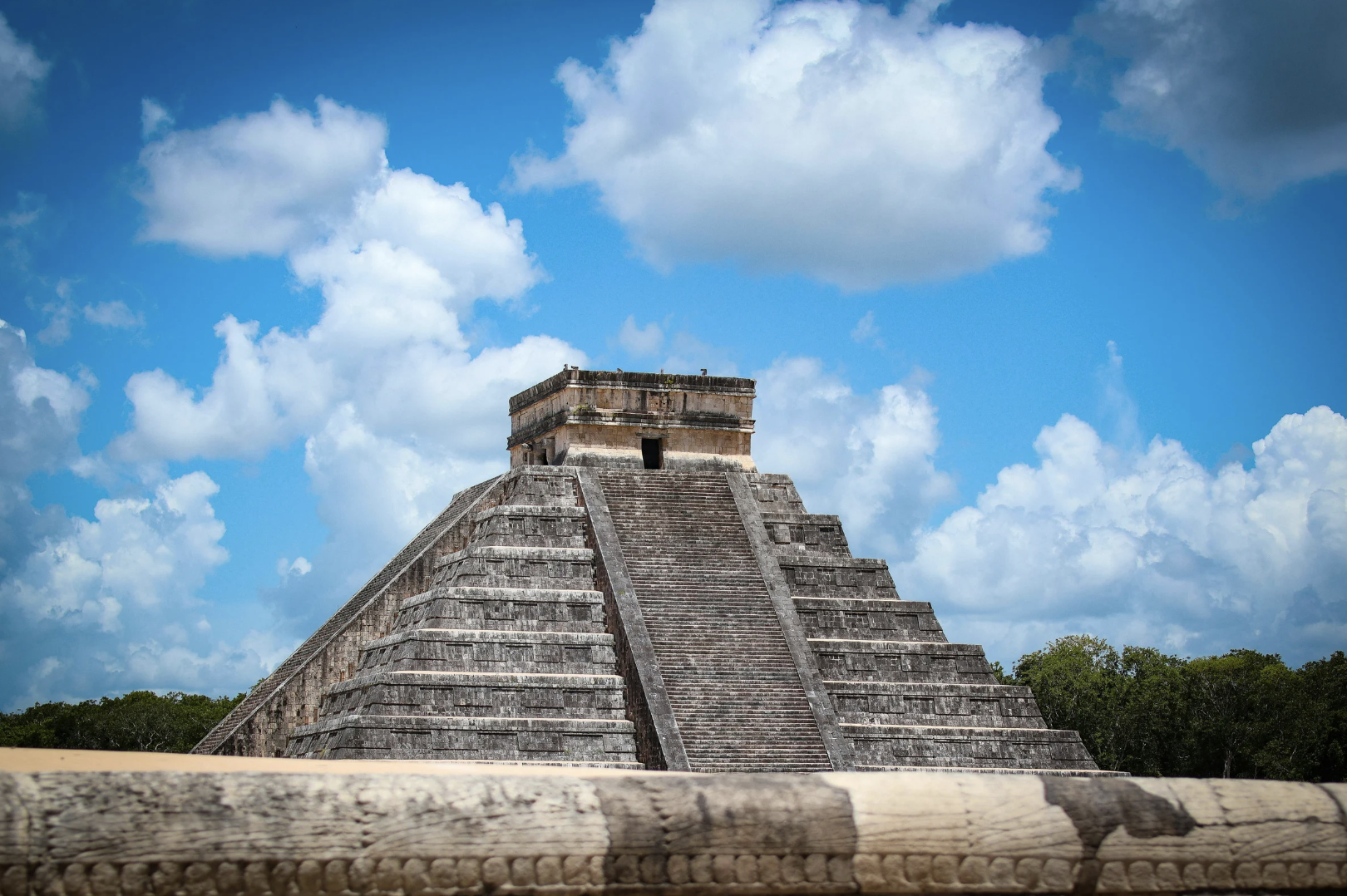 Pirámide maya antigua en un paisaje con cielo azul y nubes, rodeada de vegetación.