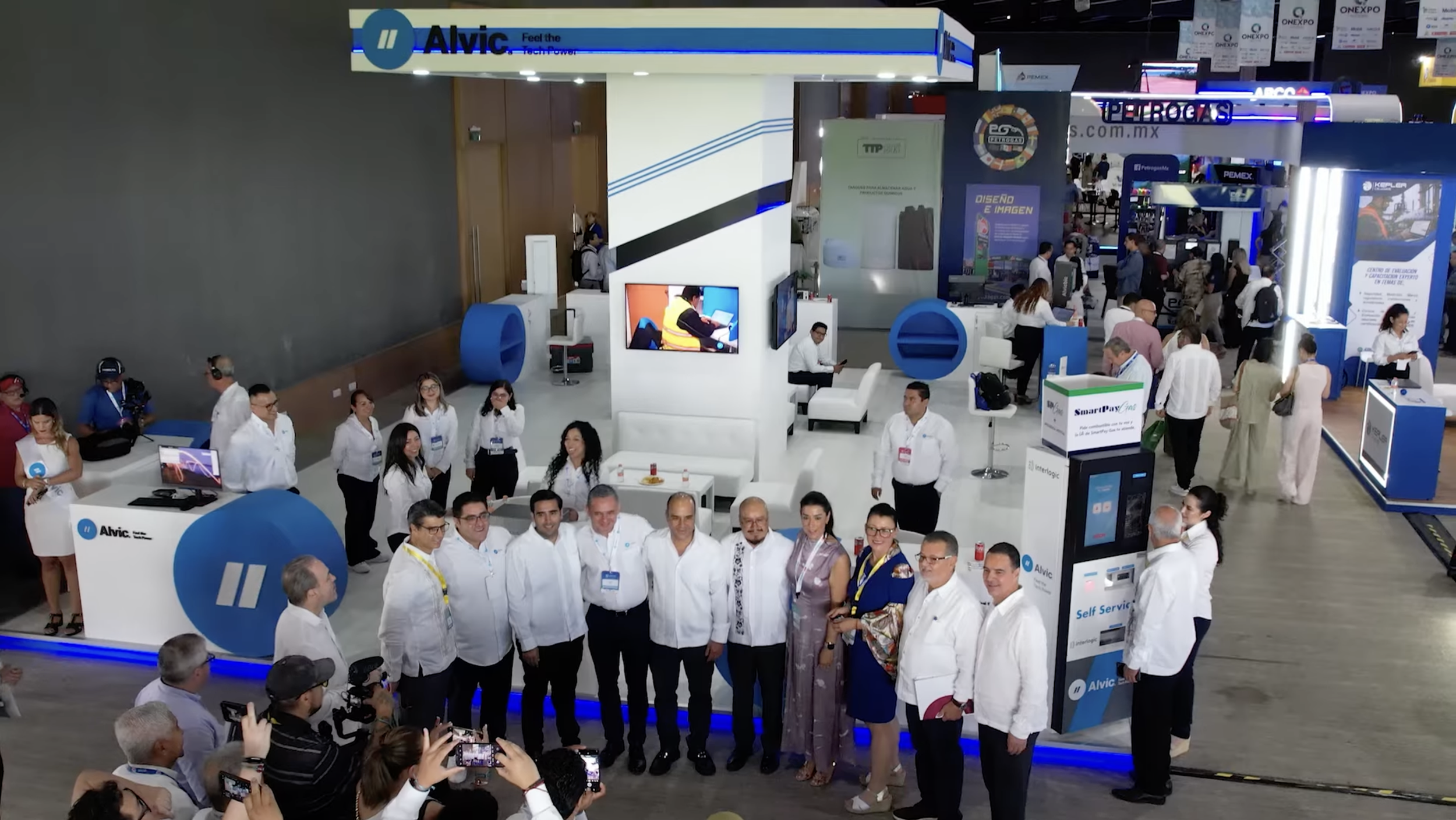 Imagen de un evento o exposición con varias personas, algunas en uniforme blanco, en un stand con logotipos de Alvic y otros, con pantallas y equipos electrónicos, en un espacio grande y concurrido.