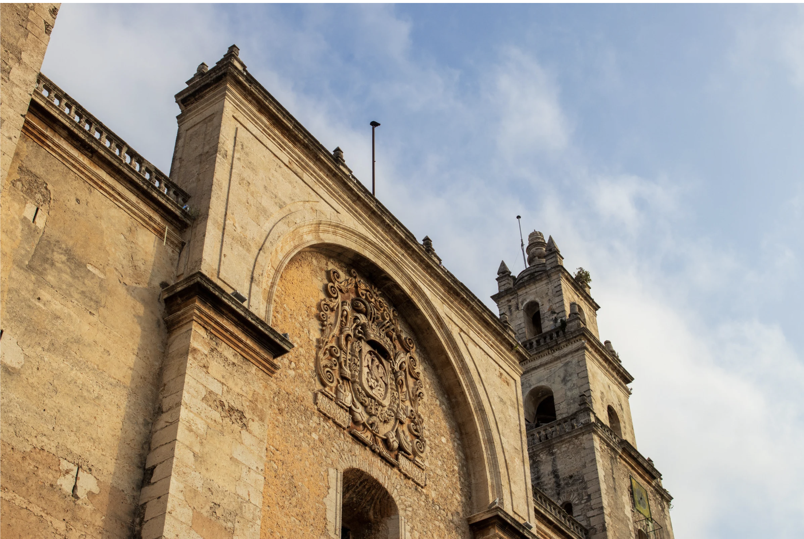 Vista de una antigua iglesia con detalles en piedra y una escultura en el centro de la fachada, con torres al fondo y un cielo con algunas nubes.