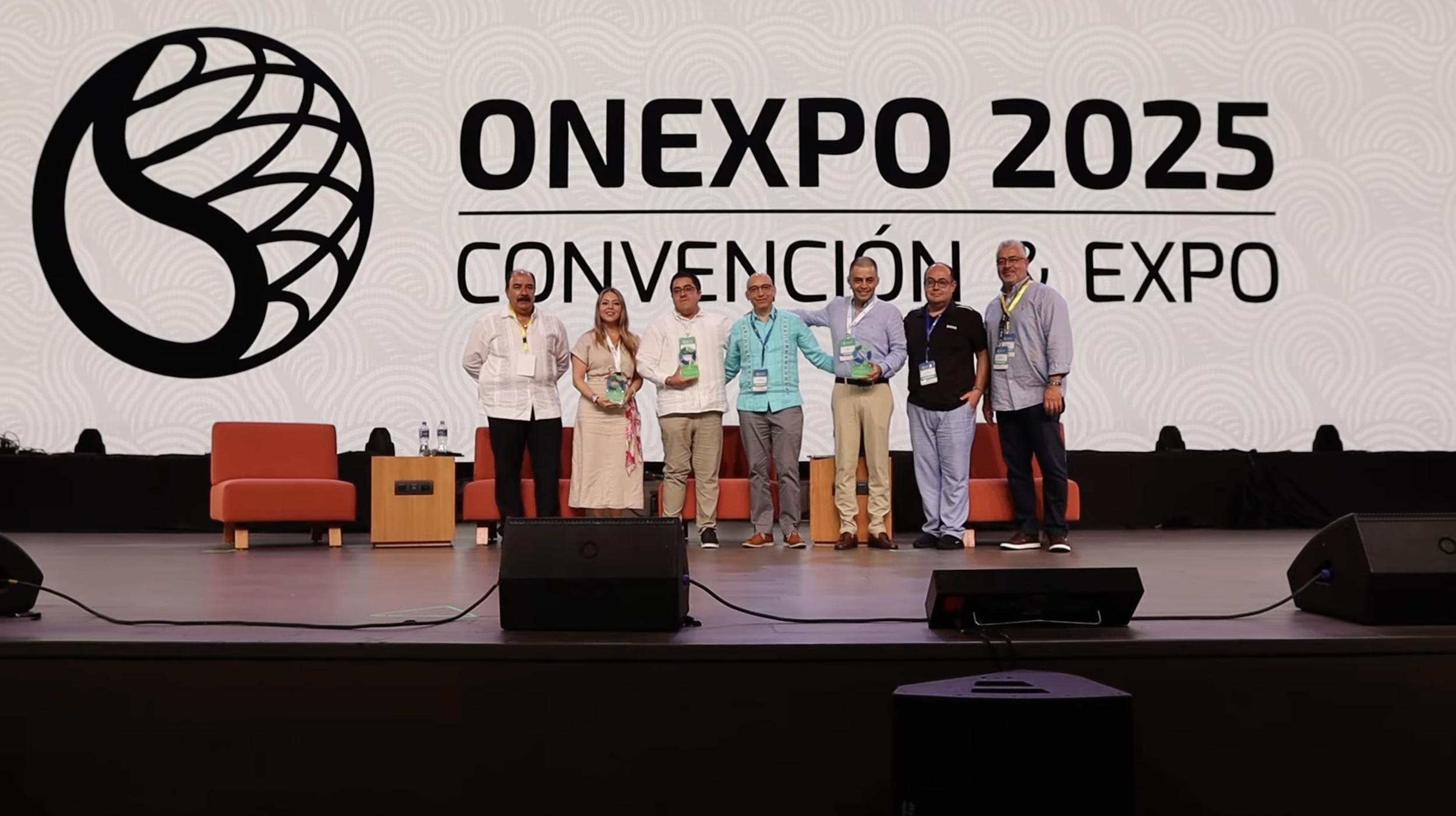 Grupo de siete personas de pie en un escenario durante la ONEXPO 2025, con un fondo grande que dice ONEXPO 2025, CONVENCIÓN Y EXPO, y un logo circular a la izquierda. La gente sostiene premios o reconocimientos y usa credenciales colgadas al cuello.