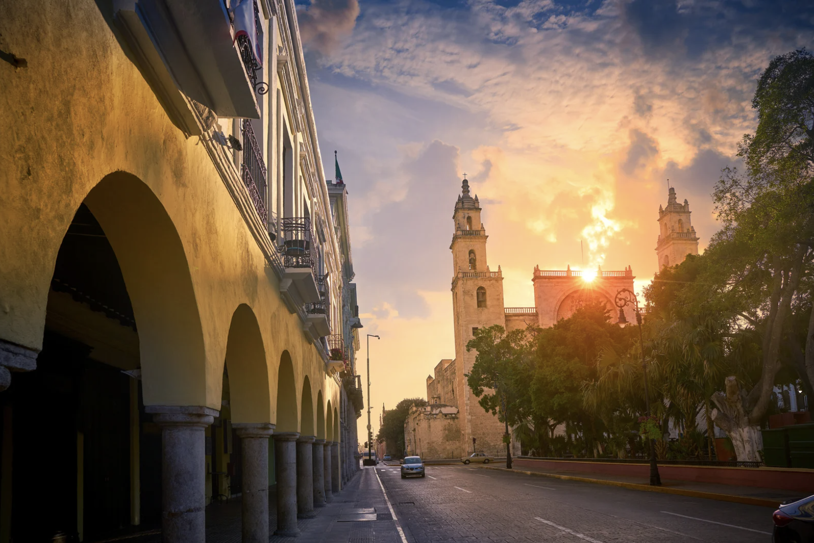 Vista de una calle con edificios coloniales, árboles y una iglesia al fondo, durante el atardecer.