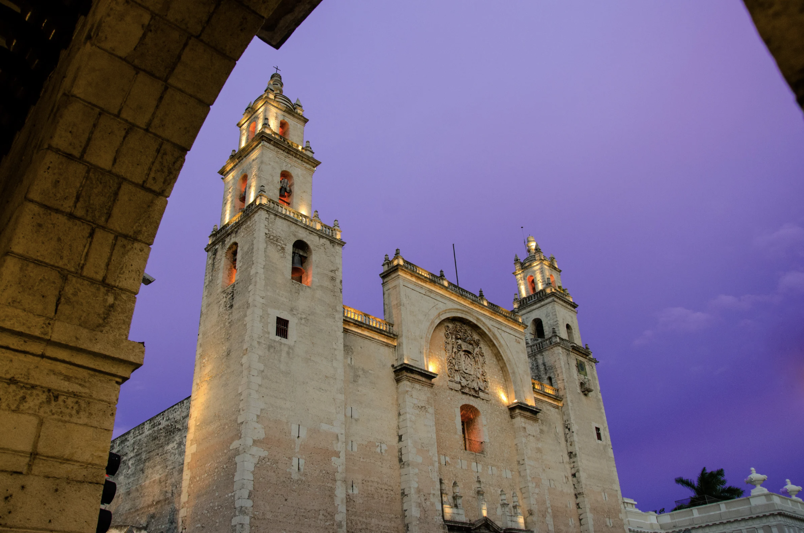 Vista de la fachada de una catedral antigua con torres altas, iluminada, en un atardecer con cielo morado y algunos edificios a su alrededor.