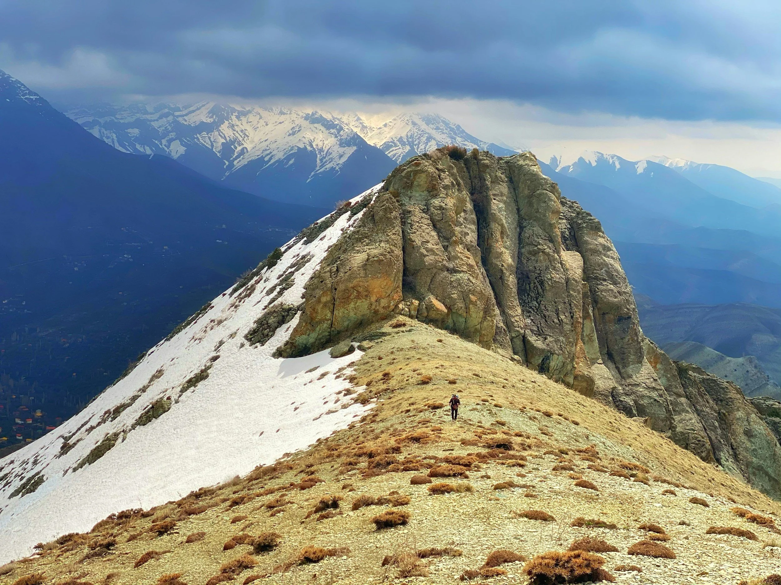 Image of reaching the peak or pinnacle. A hiker walking on a mountain ridge with snow patches and large rock formations, with snow-capped peaks and a cloudy sky in the background.