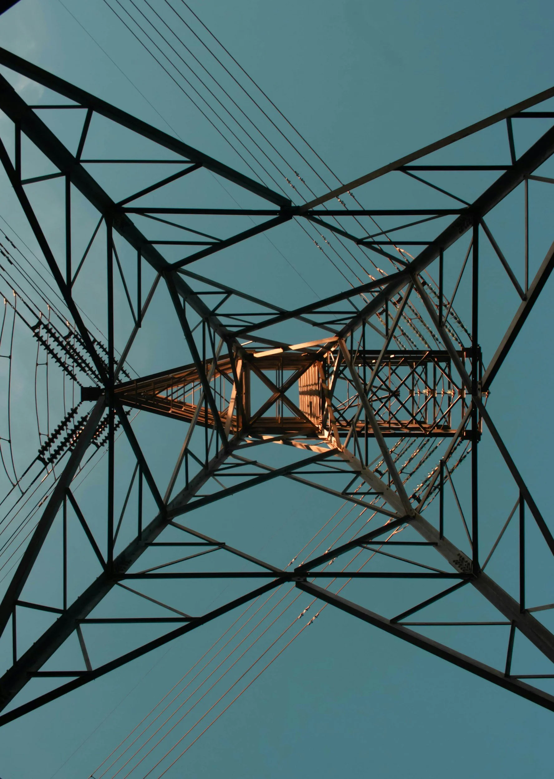 Looking up at a tall electrical transmission tower with wires extending into the sky, illuminated by sunlight.
