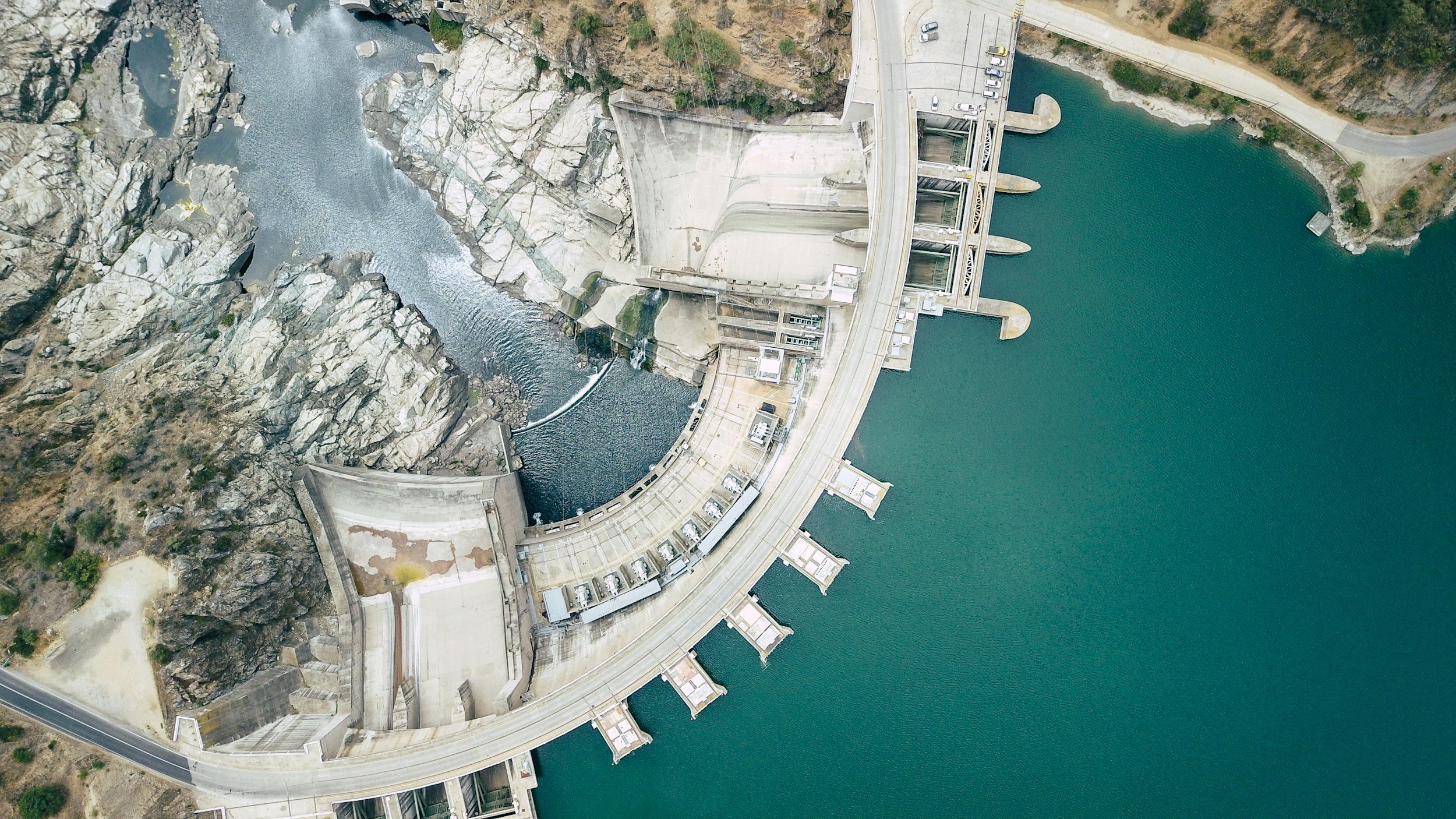 Aerial view of a hydroelectric dam with spillways, water reservoir, and surrounding rocky terrain.