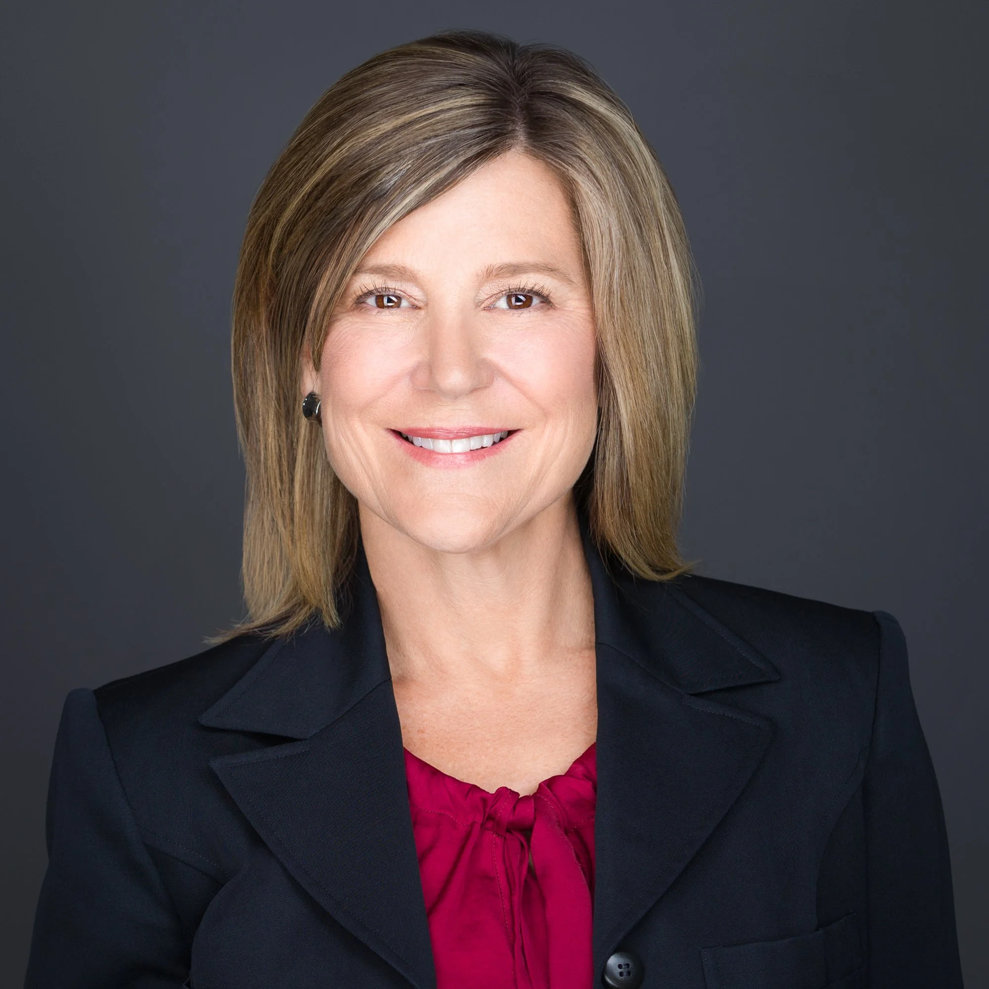 The Principal of Harness Strategy. A professional woman with shoulder-length blonde hair smiling, wearing a black blazer and a red blouse, against a dark gray background.