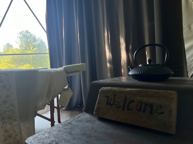 A cozy interior with a mattress on a wooden bed frame, dark curtains, a table with a wooden "Welcome" sign, and a black kettle on a dark surface, with a large window showing trees outside.