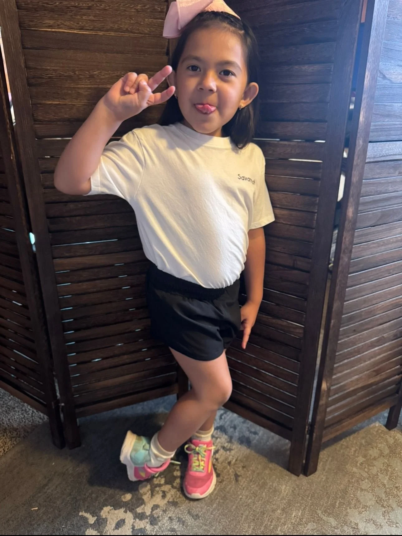 A young girl making a silly face and holding up a peace sign with her right hand. She is standing between two wooden room dividers, wearing a light-colored t-shirt, dark shorts, colorful sneakers, and a pink bow in her hair.
