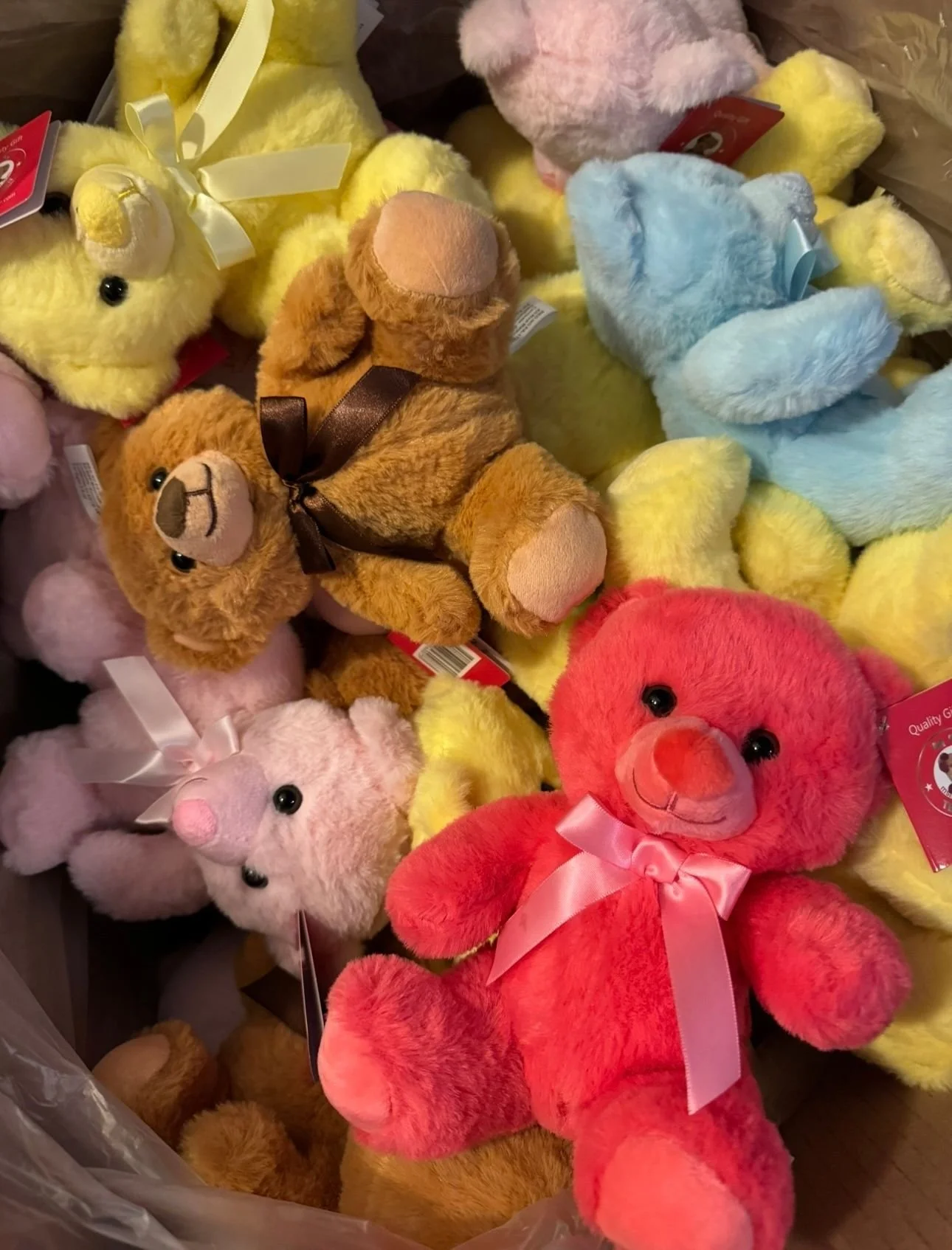 A collection of colorful plush teddy bears in various colors including pink, red, yellow, blue, brown, and beige, with some wearing ribbons or bows. Savvy's Stuffies.