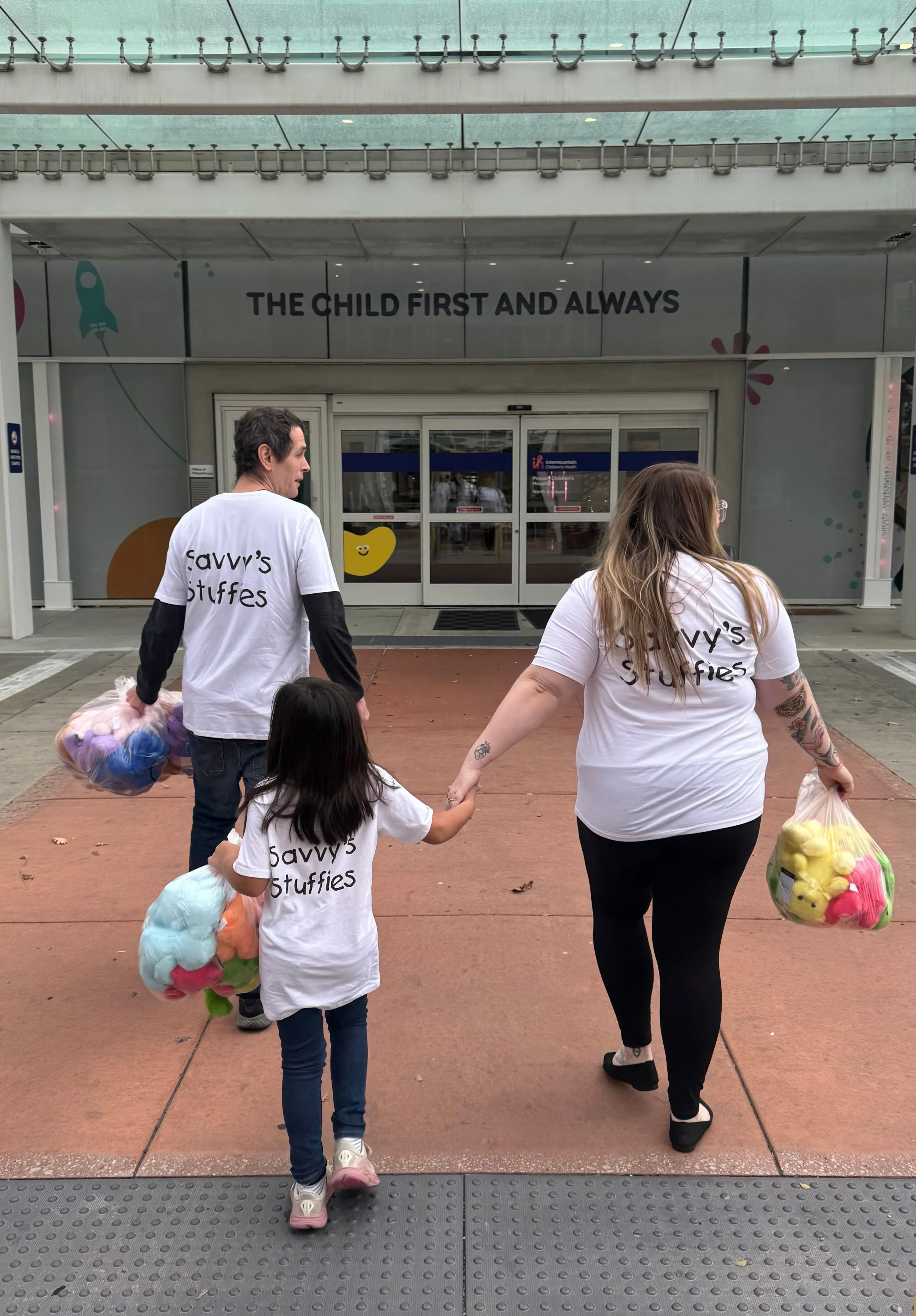 A family of three walking into Primary Children's Hospital carrying teddy bears, with T-shirts that say "Savvy's Stuffies." They are exiting through a glass entrance, and above the door is a sign that says "The Child First and Always."
