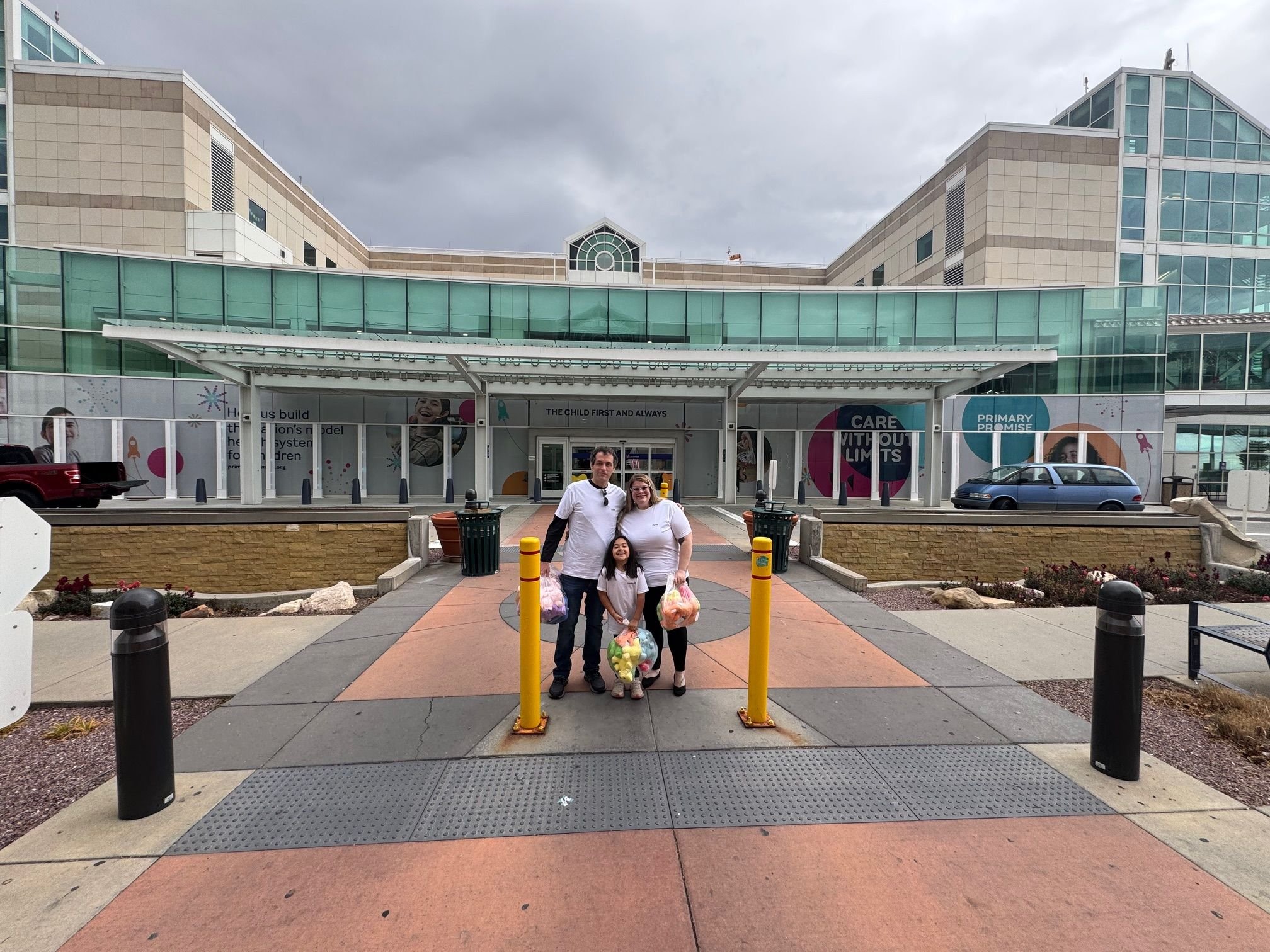 Family of three standing outside primary childrens hospital building with teddy bear donations, on a cloudy day. Savvy's Stuffies.