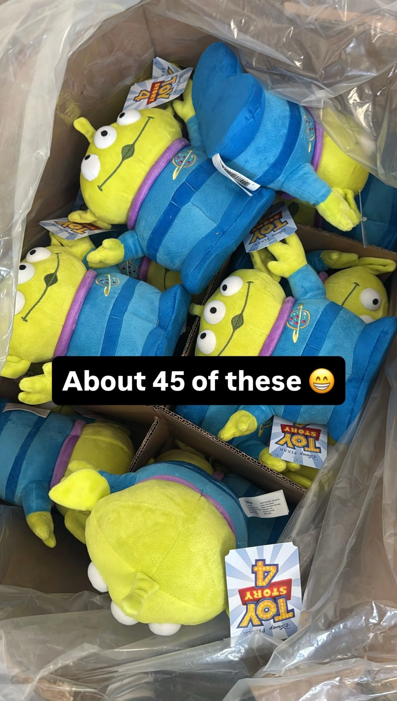 A large quantity of Toy Story plush toys, featuring aliens with three eyes and blue spacesuits, popping out of a cardboard box lined with a plastic bag, with a black overlay text stating 'About 45 of these' with a smiling emoji.