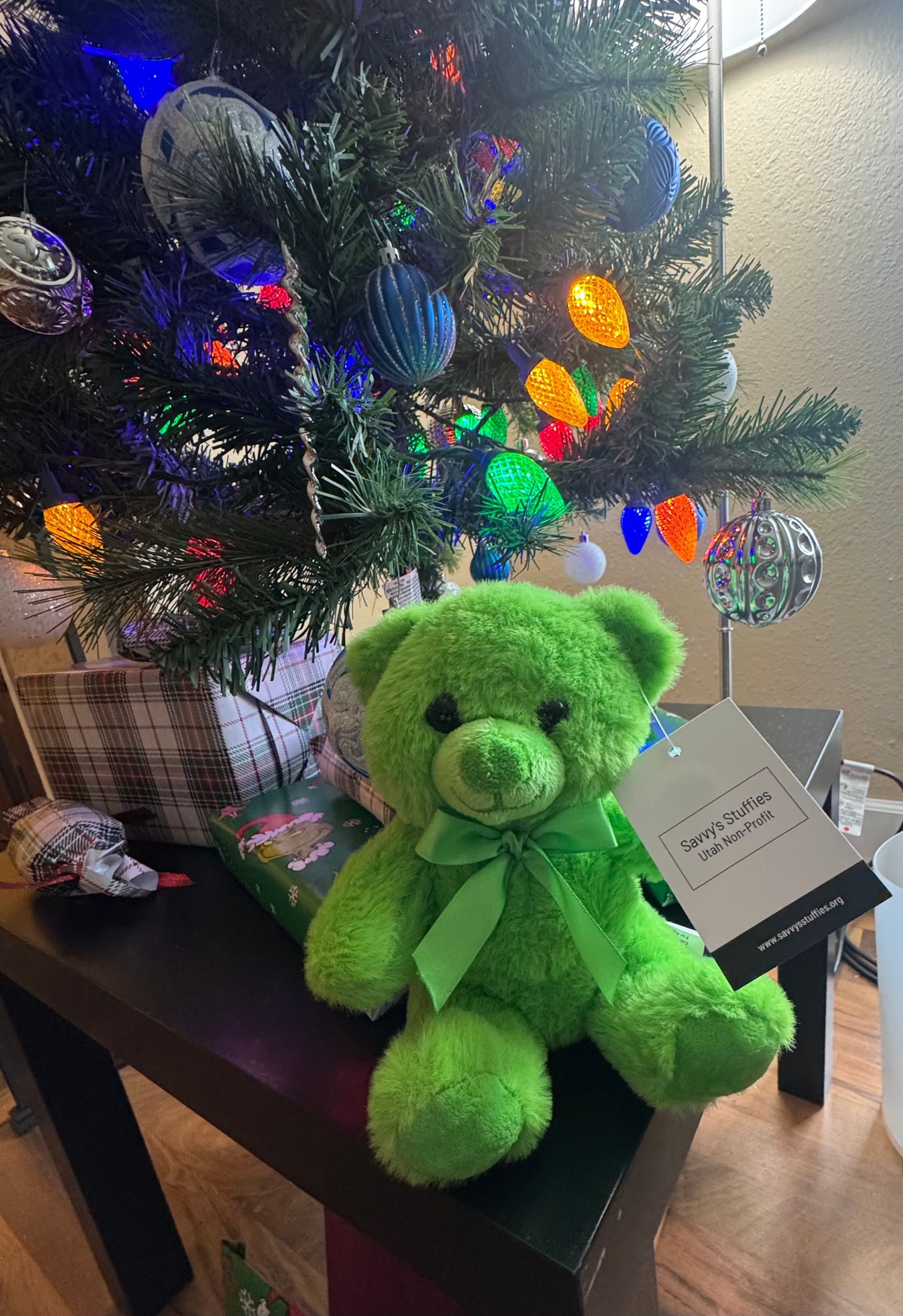 Green teddy bear with a green ribbon sitting in front of a decorated Christmas tree with colorful lights and ornaments, surrounded by wrapped presents. Savvy's Stuffies