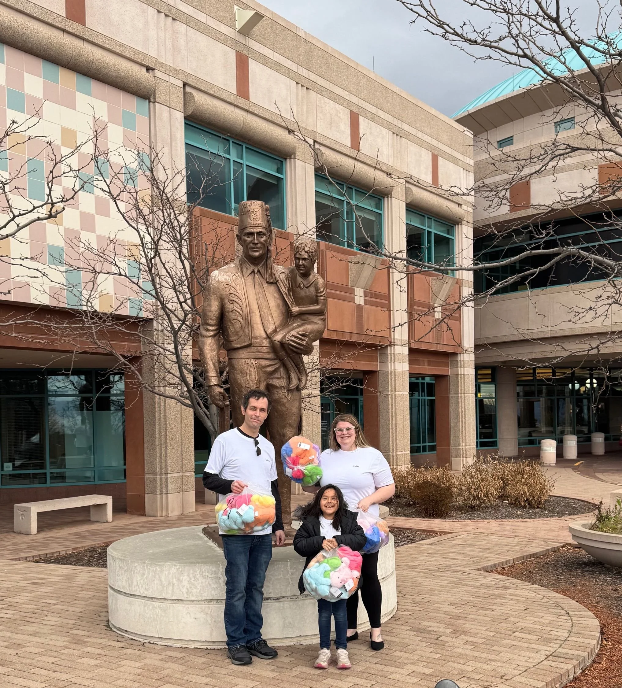 two adults and a child, standing in front of a large bronze statue of a man holding a child. holding bags of colorful stuffed toys. outside of a modern building with large windows and leafless trees. Shriners childrens hospital. Savvy's Stuffies.