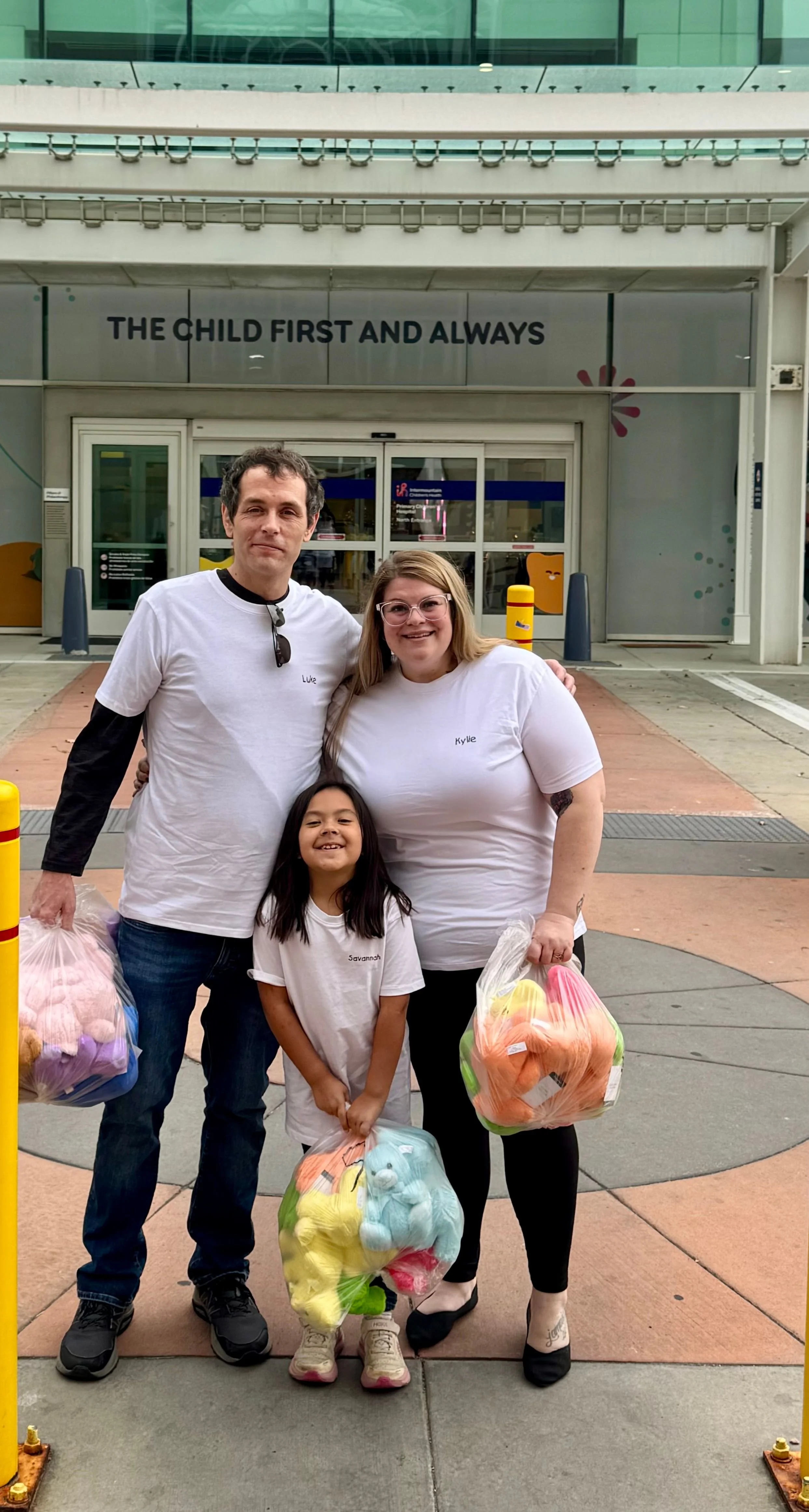 A family of three standing outside a hospital with the sign 'The Child First and Always.' The man, wearing a white T-shirt and jeans, holds a bag of toys. The woman, also in a white T-shirt, has glasses and blonde hair. The young girl, smiling, has l