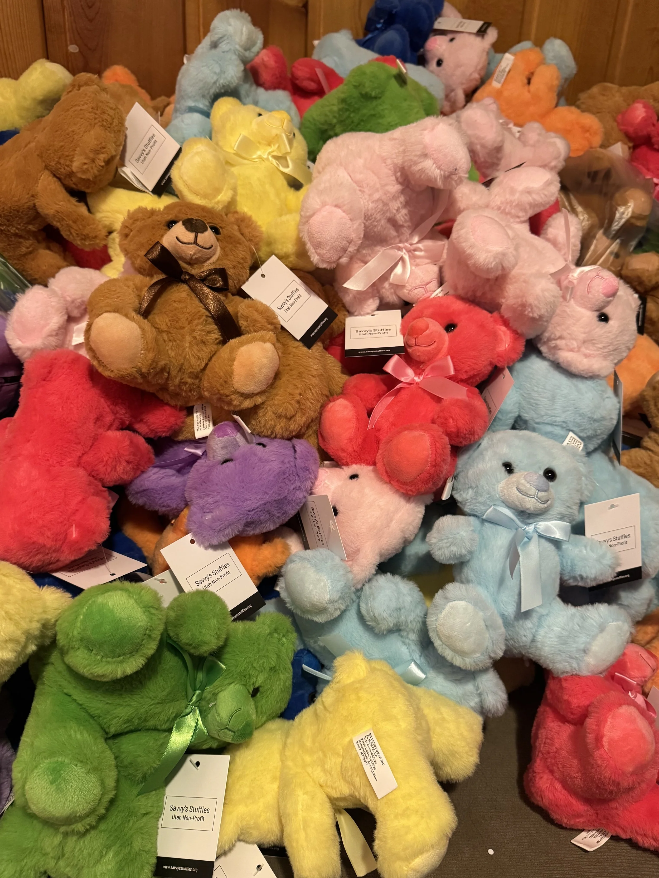A colorful assortment of plush teddy bears in various colors including brown, yellow, green, pink, red, purple, blue, and orange, with some bears having ribbons around their necks, displayed on a surface. Savvy's Stuffies.