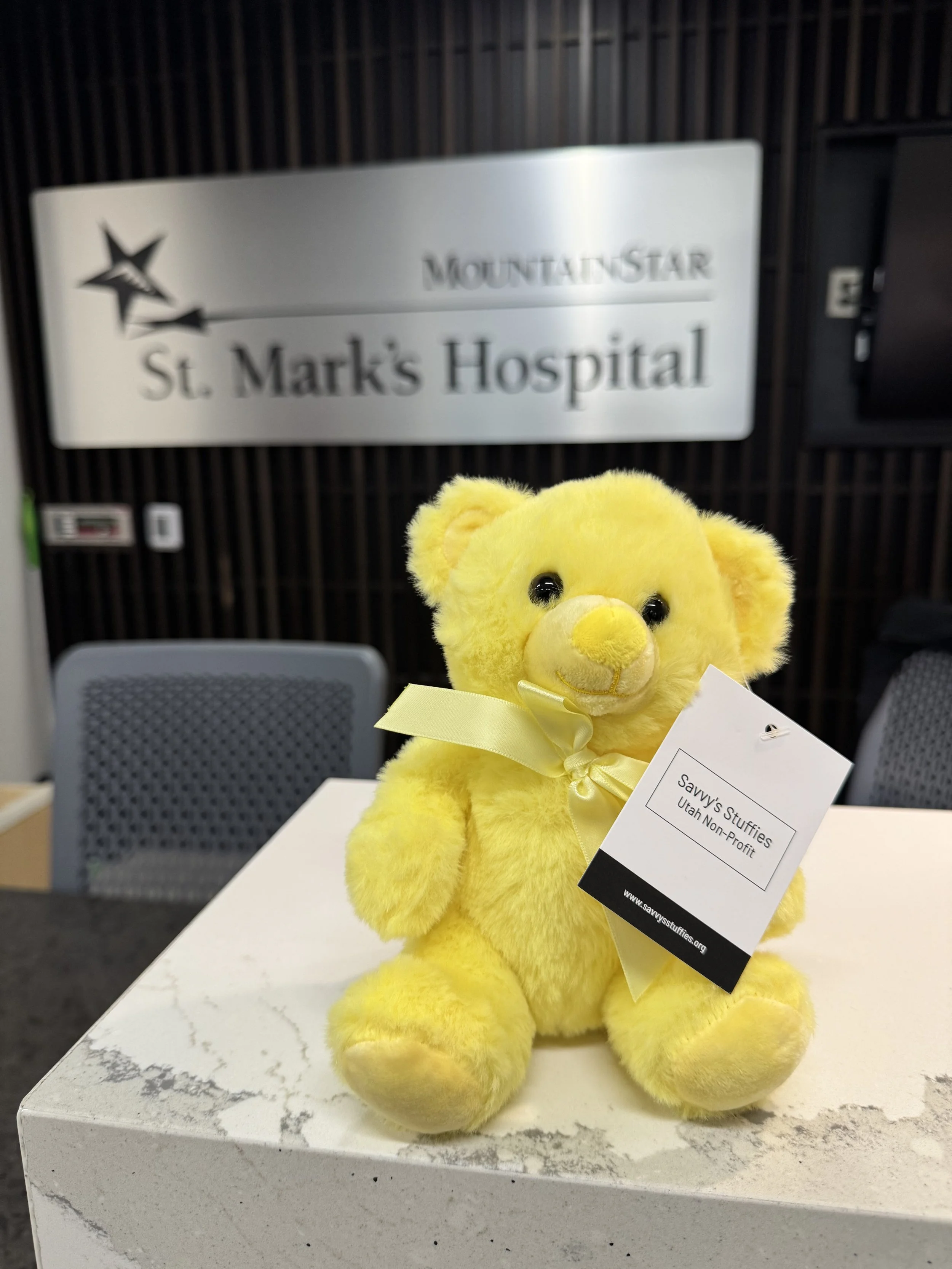 Yellow plush teddy bear with a yellow ribbon sitting on a white surface in front of a hospital sign that reads "Mountainside St. Mark's Hospital."
