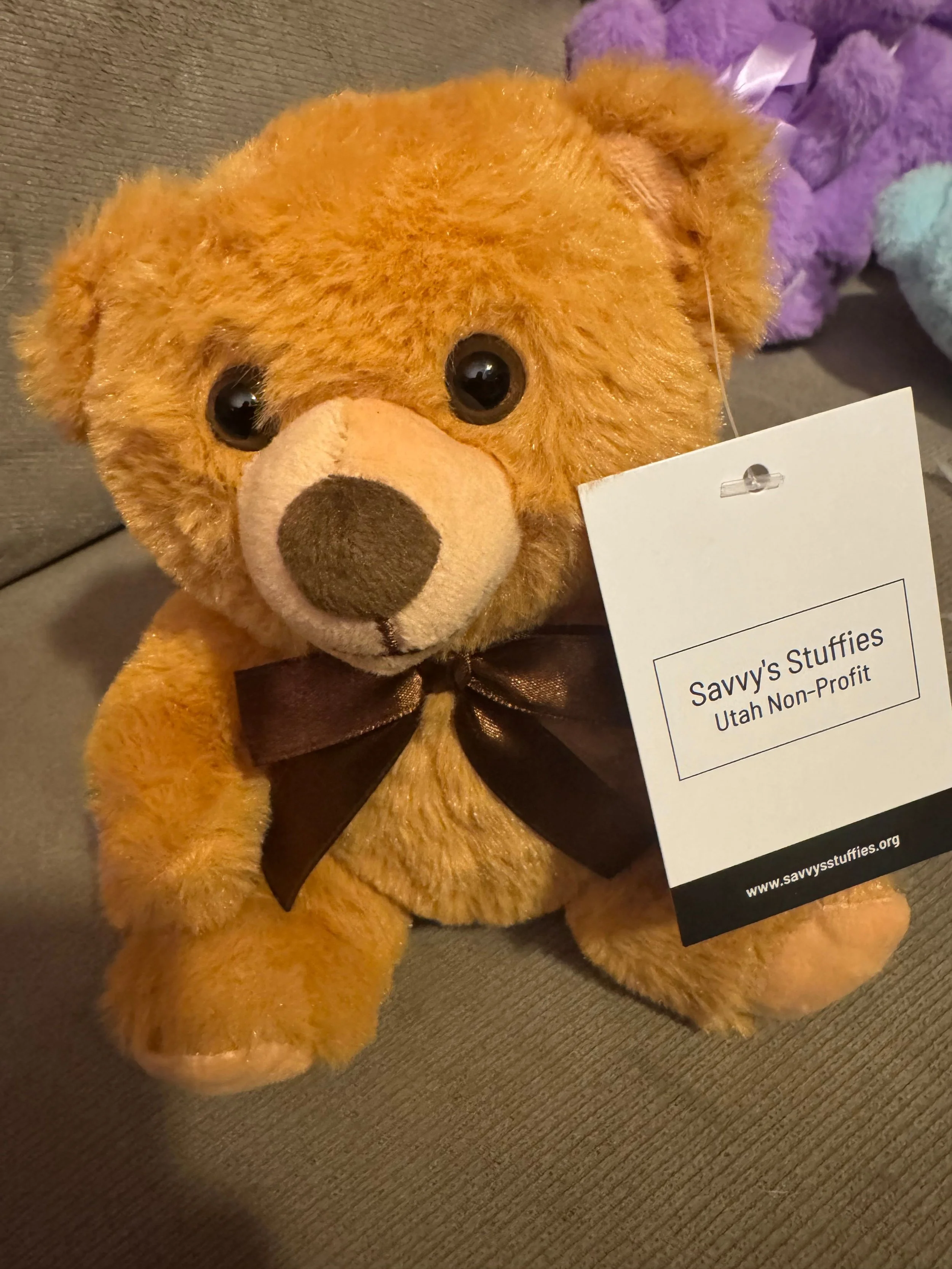 A plush teddy bear with brown fur, large black eyes, a beige nose, and a dark brown ribbon tied around its neck, with a tag from Savvy's Stuffies, Utah Non-Profit.
