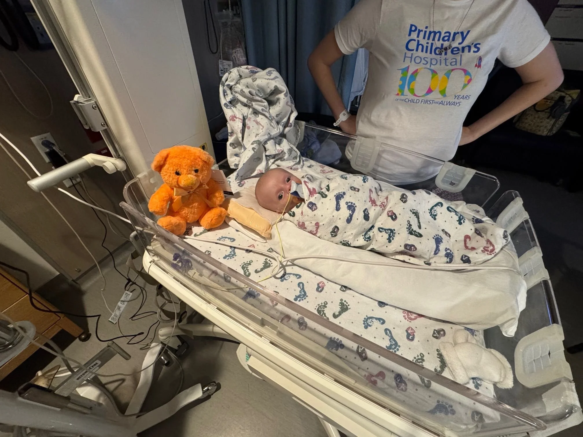 A baby lying in a hospital bassinet with a nasal tube, wrapped in a colorful footed blanket, next to an orange teddy bear plush toy. A person, wearing a shirt that reads "Primary Children's Hospital 100 Years," stands beside the bassinet.