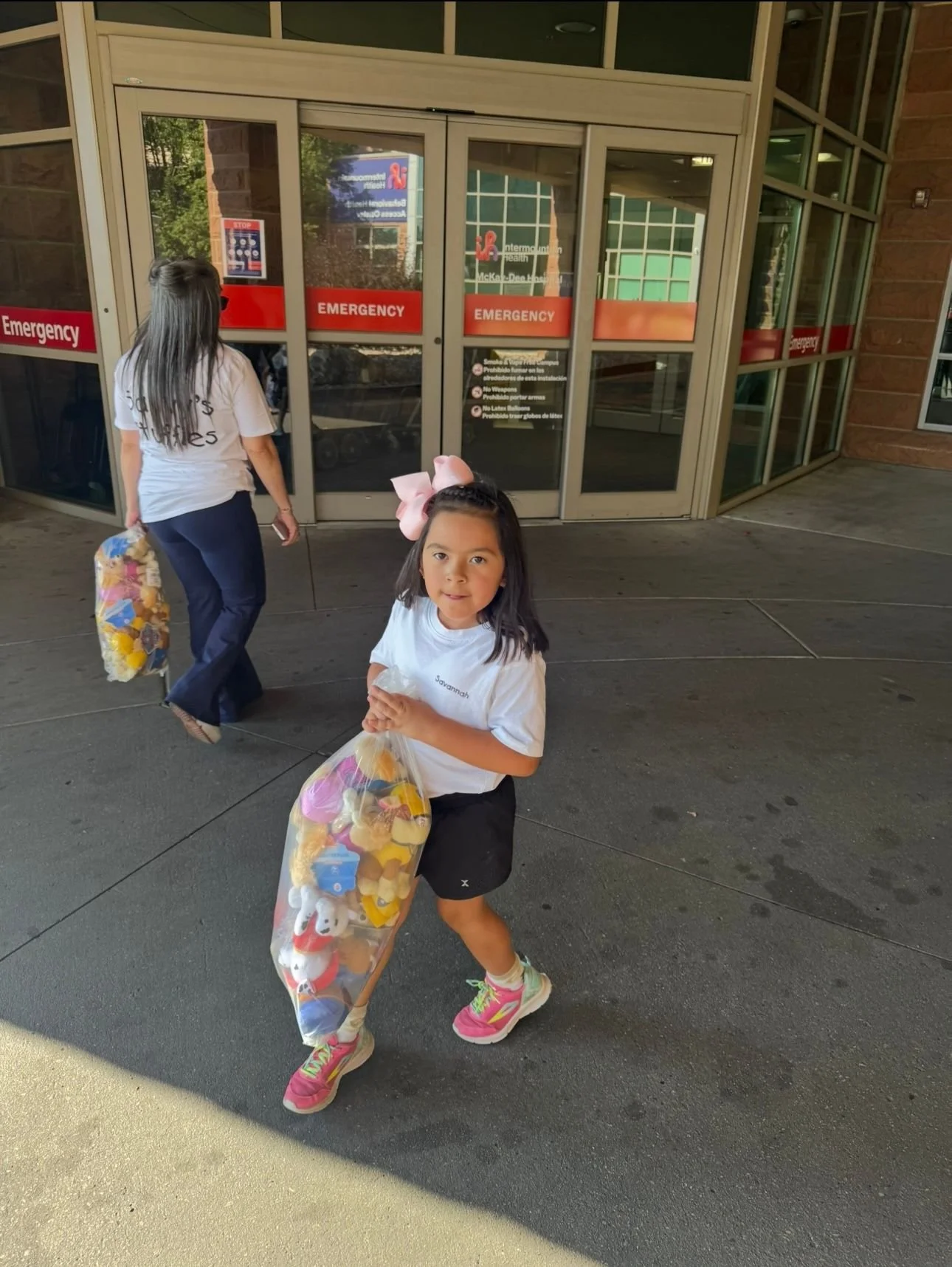 A young girl with a pink bow in her hair, holding a large bag of plush toys, standing outside McKay Dee Hospital entrance. An adult woman is walking away in the background carrying a bag of toys. Savvy's Stuffies.
