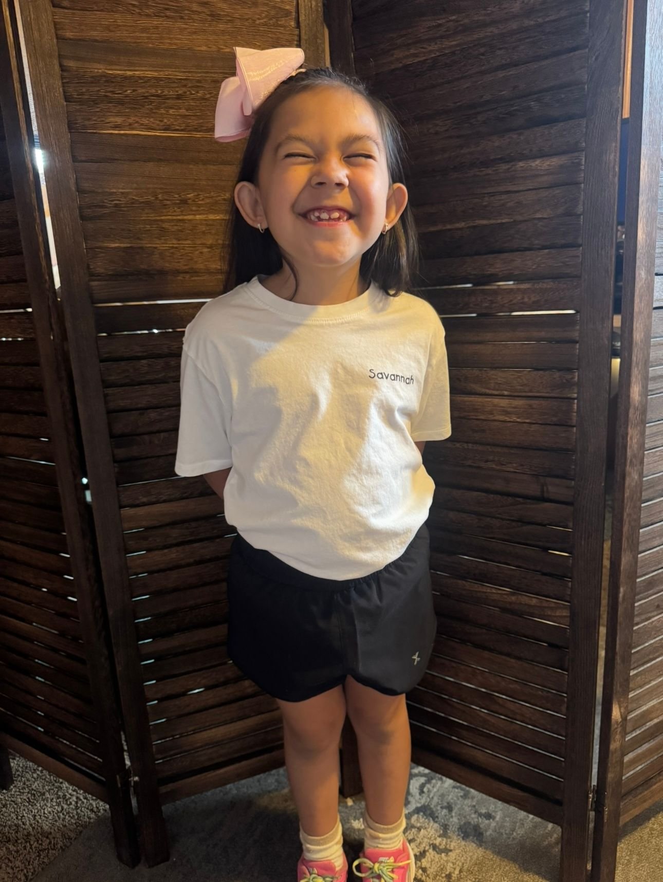 A young girl with a pink bow in her hair, smiling broadly with eyes closed, standing indoors in front of a wooden divider, wearing a white T-shirt with inky text and black shorts, pink shoes, and yellow socks.