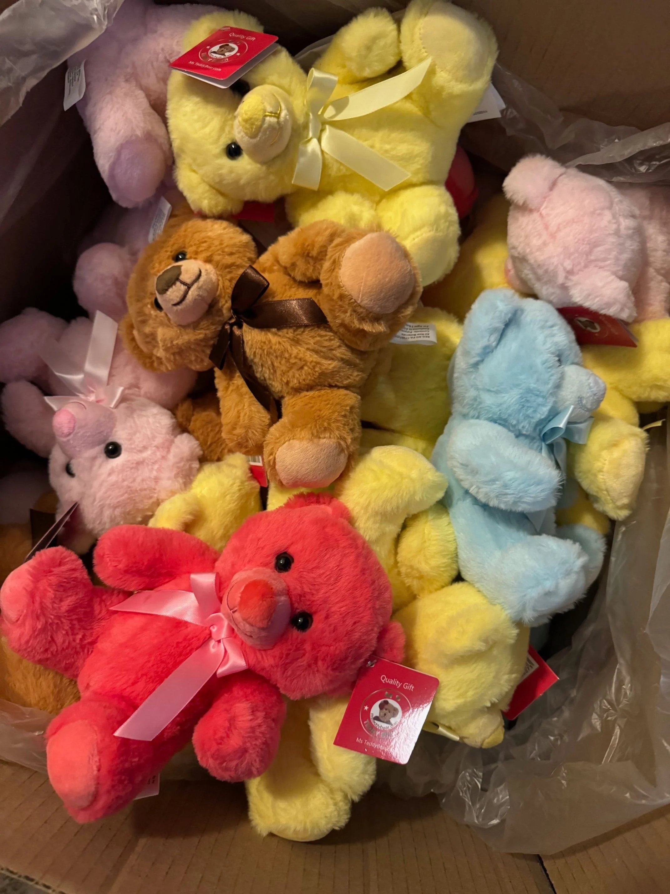 A box filled with colorful plush teddy bears of various colors including yellow, pink, blue, brown, and red. Some bears have ribbons tied around their necks, and a few have tags attached, indicating they are gifts. Savvy's Stuffies.