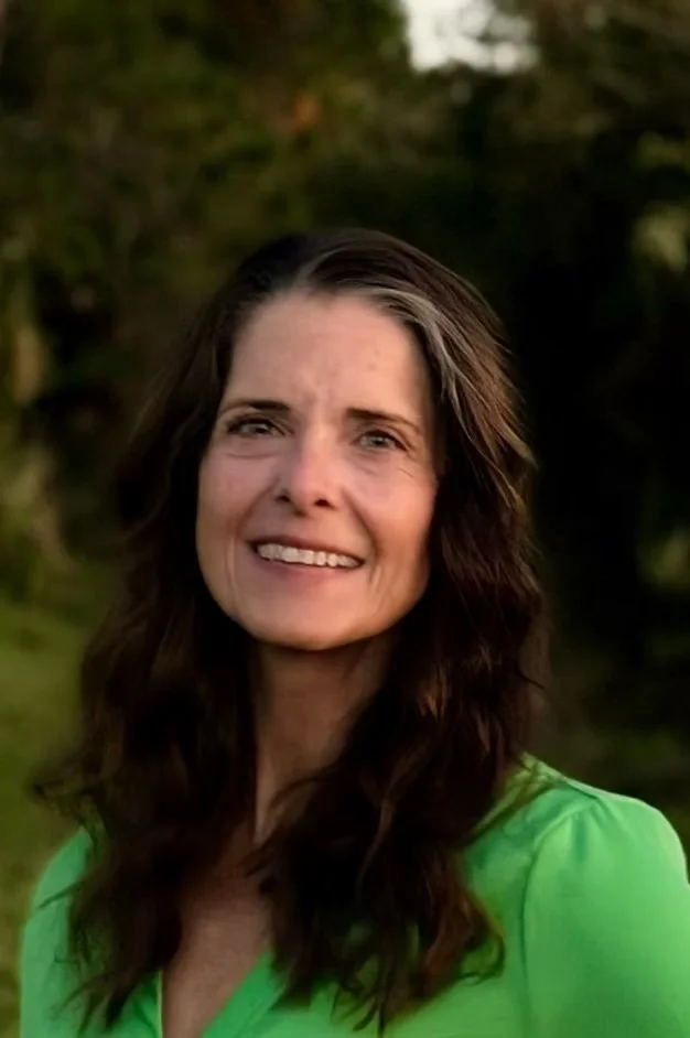 Portrait of a woman with long wavy brown hair wearing a green shirt, outdoors with blurred greenery background.