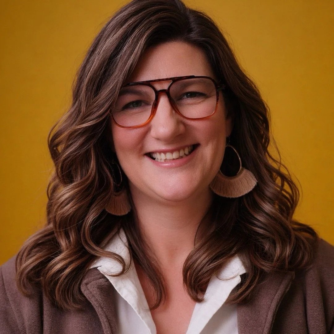 A woman with wavy brown hair wearing glasses, hoop earrings, a white shirt, and a brown cardigan, smiling against a yellow background.