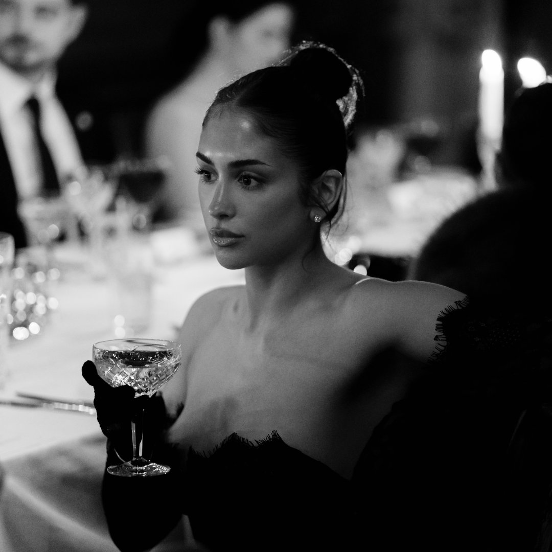 A woman with dark hair styled in a bun, wearing elegant earrings and black gloves, sitting at a table holding a glass of champagne.