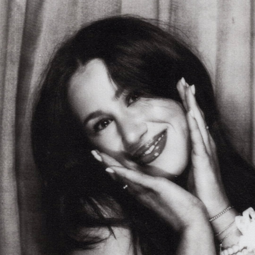 Black and white photo of a woman smiling, with her hands gently touching her face, long dark hair, and a curtain in the background.
