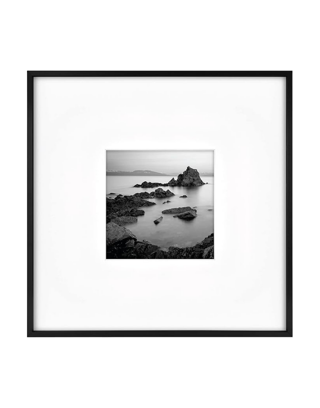 Black and white photograph of a rocky seascape with a large rock formation in the water, distant landmass in the background, and calm water with reflections.