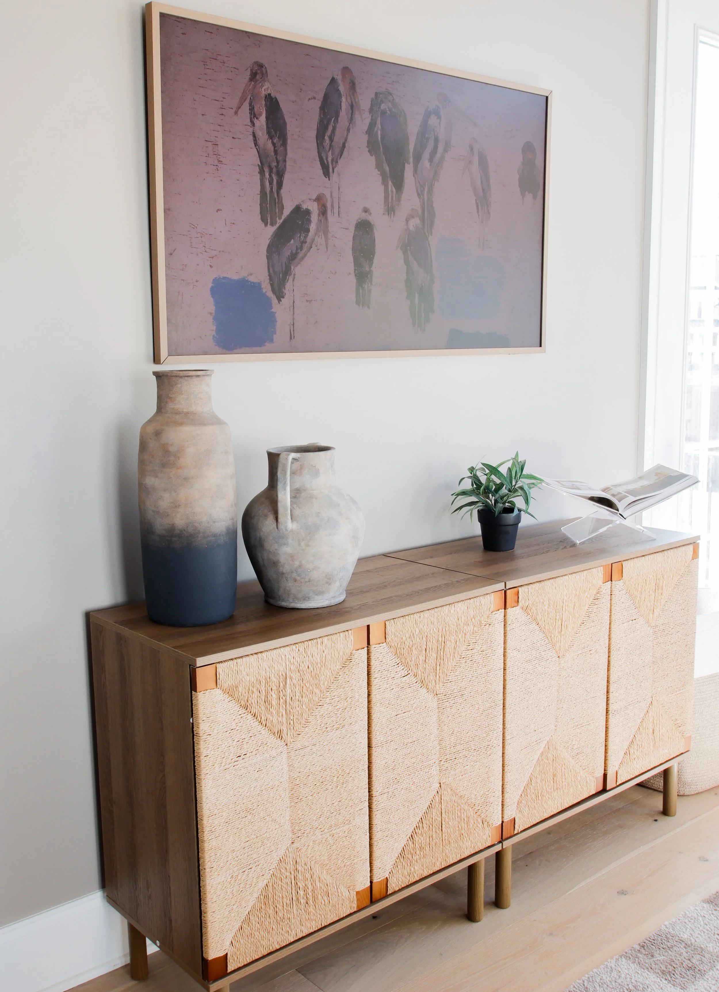 A wooden sideboard with woven cabinet doors, decorated with two vases, a small potted plant, and an open magazine, with a framed artwork of abstract birds hanging above.