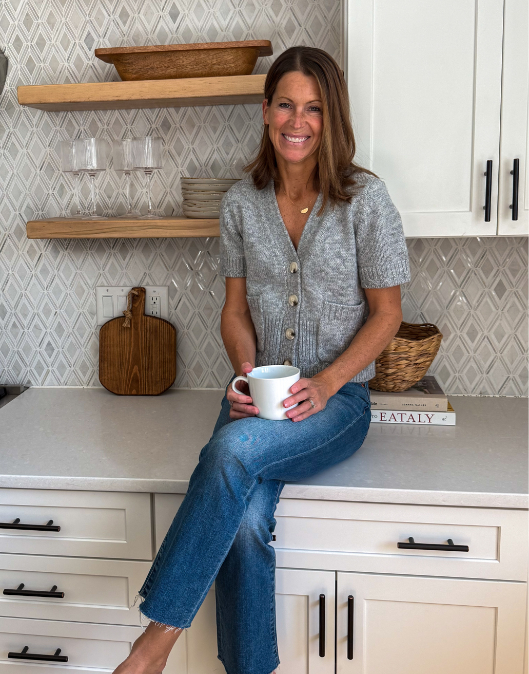 A woman sitting on a kitchen countertop, holding a white mug, smiling at the camera. She has shoulder-length brown hair and is wearing a gray short-sleeve cardigan with buttons and blue jeans.