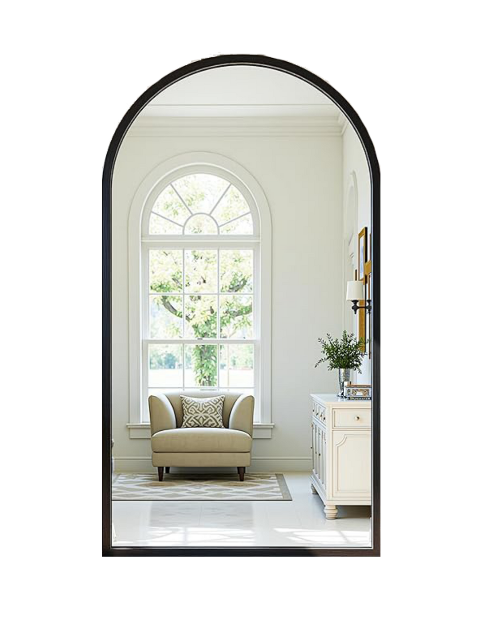 View of a cozy corner with a cream-colored armchair, decorative pillow, a white side table with a vase of greenery and a lamp, large arched window with greenery outside, and a framed picture on the wall.