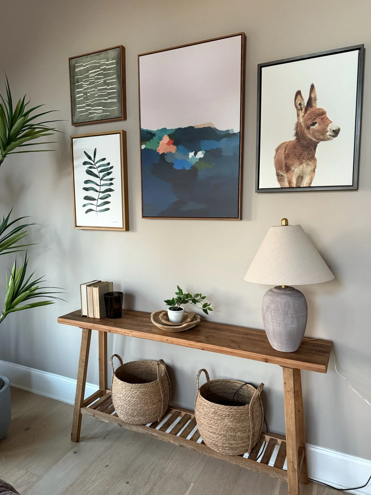 Decorative living room wall with five framed art pieces, a wooden console table with a table lamp, potted plant, books, and two woven baskets underneath.