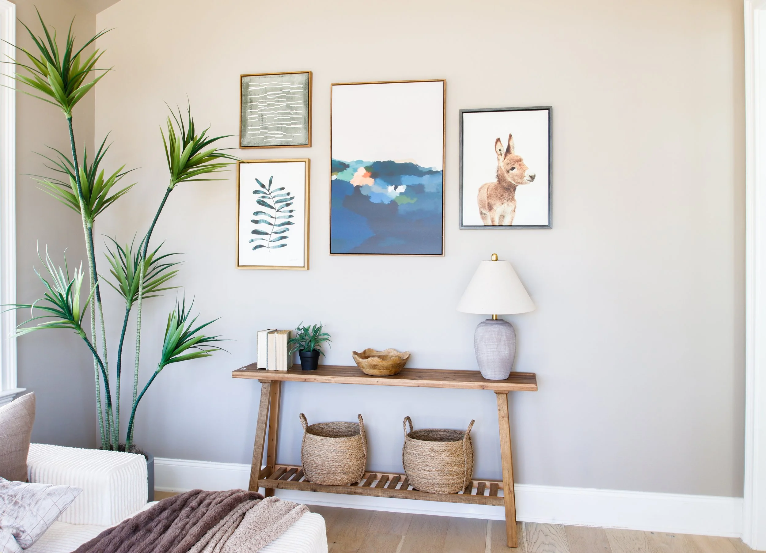 A living room wall decorated with five framed art pieces, including abstract artwork, a leaf print, a landscape, and a donkey illustration. There is a wooden console table with books, a potted plant, a wooden bowl, and a table lamp. A large potted pl