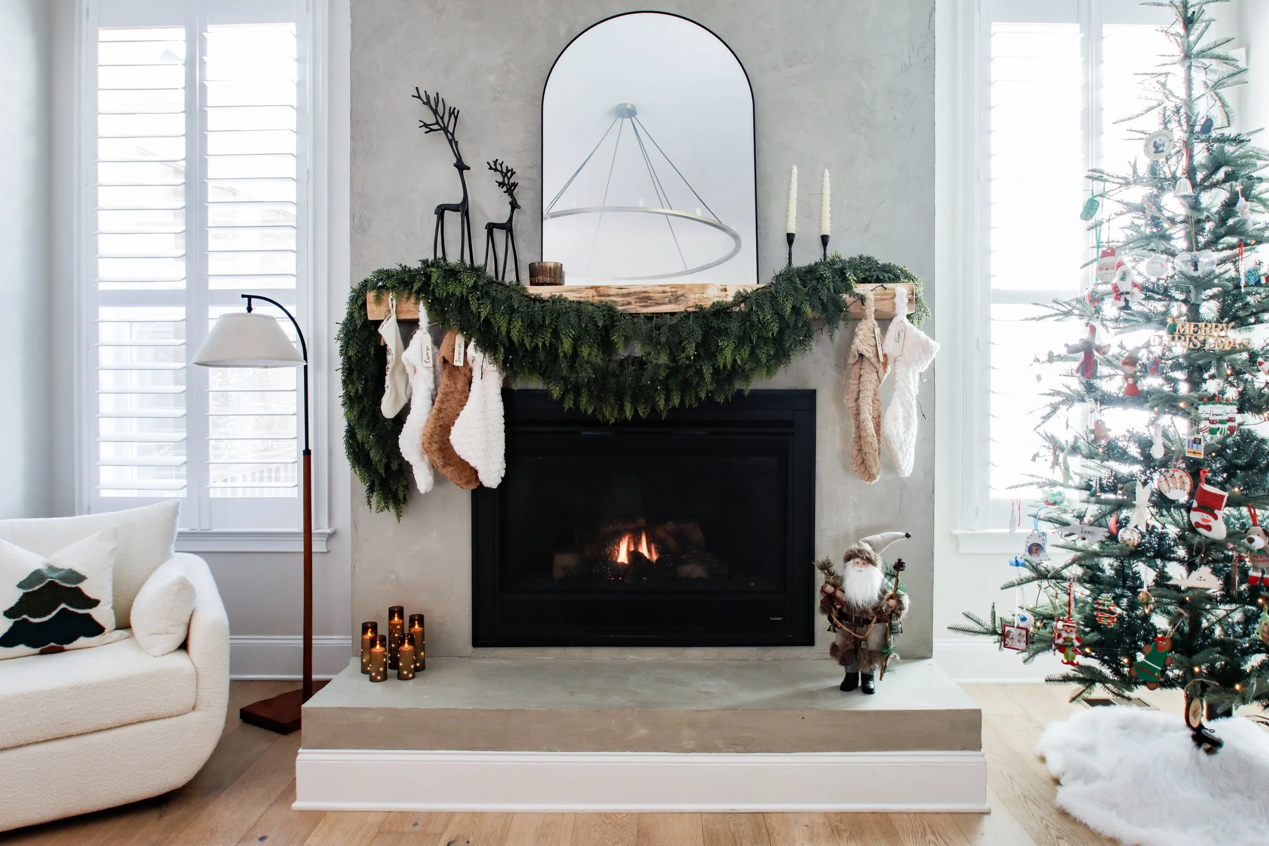 Living room fireplace decorated for Christmas with stockings, a green garland, and a mirror above the mantle. To the right, a Christmas tree with ornaments, lights, and a plush Santa figurine nearby.