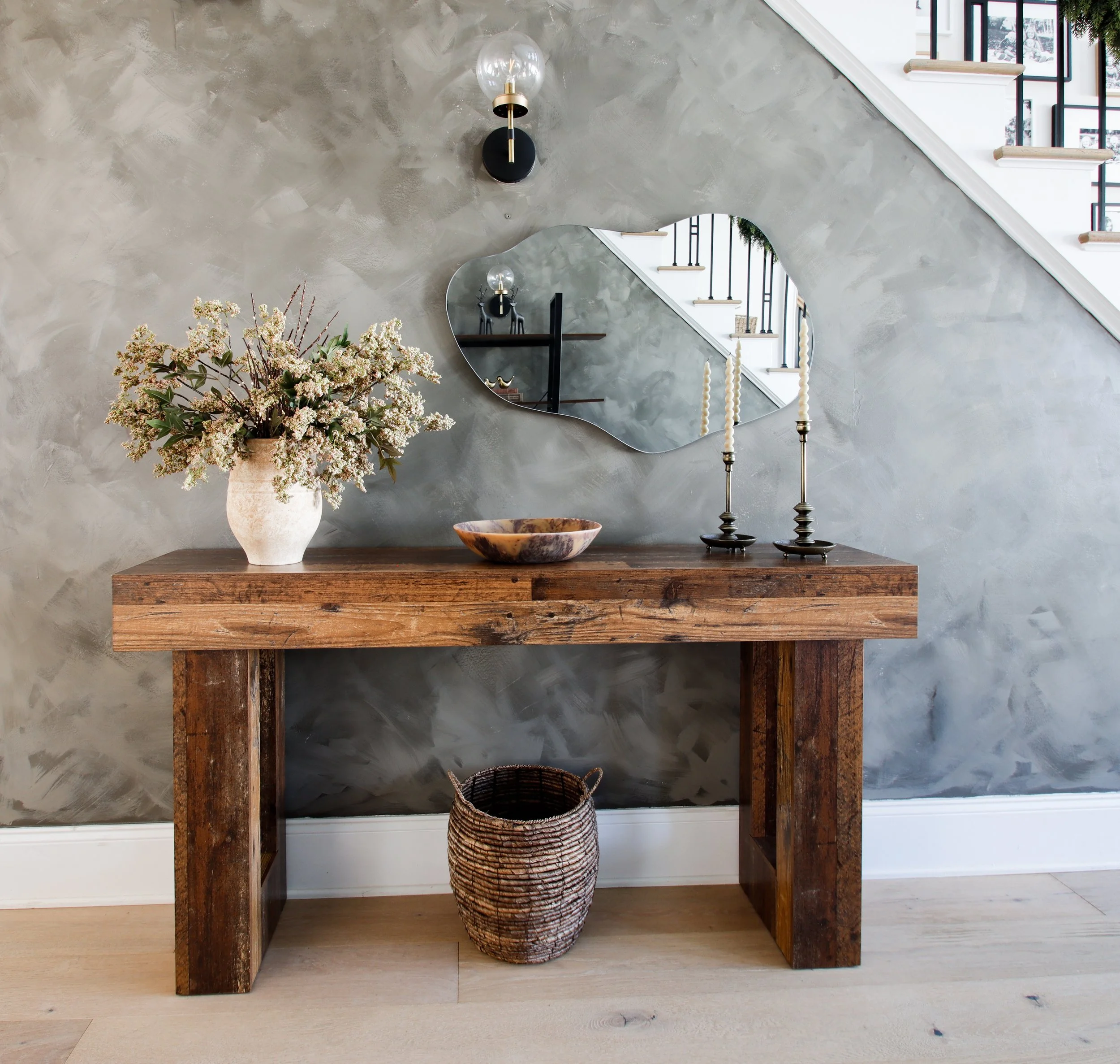 A wooden console table with a vase of flowers, a decorative bowl, and two candlesticks on it, against a textured gray wall with a mirror and a wall sconce, and a staircase reflected in the mirror.