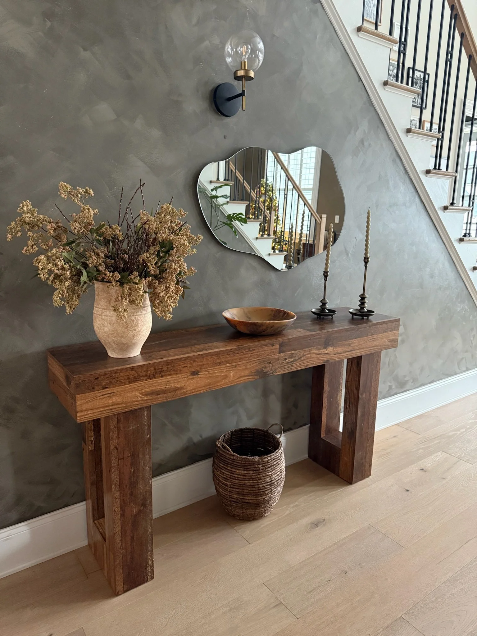 A wooden console table against a gray textured wall with a heart-shaped mirror, a large ceramic vase with dried flowers, a wooden bowl, and two tall candlesticks, with a staircase in the background.