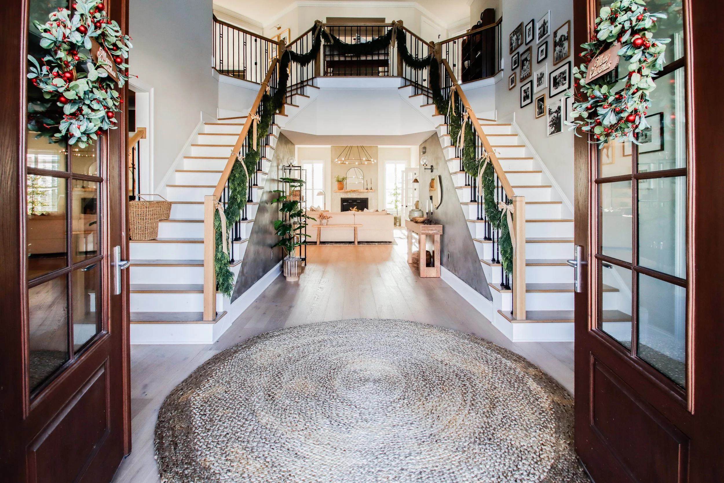 View of a decorated home interior entrance with double staircase, photo of a cozy living room area, walls adorned with framed pictures, holiday wreaths on the door, and warm sunlight