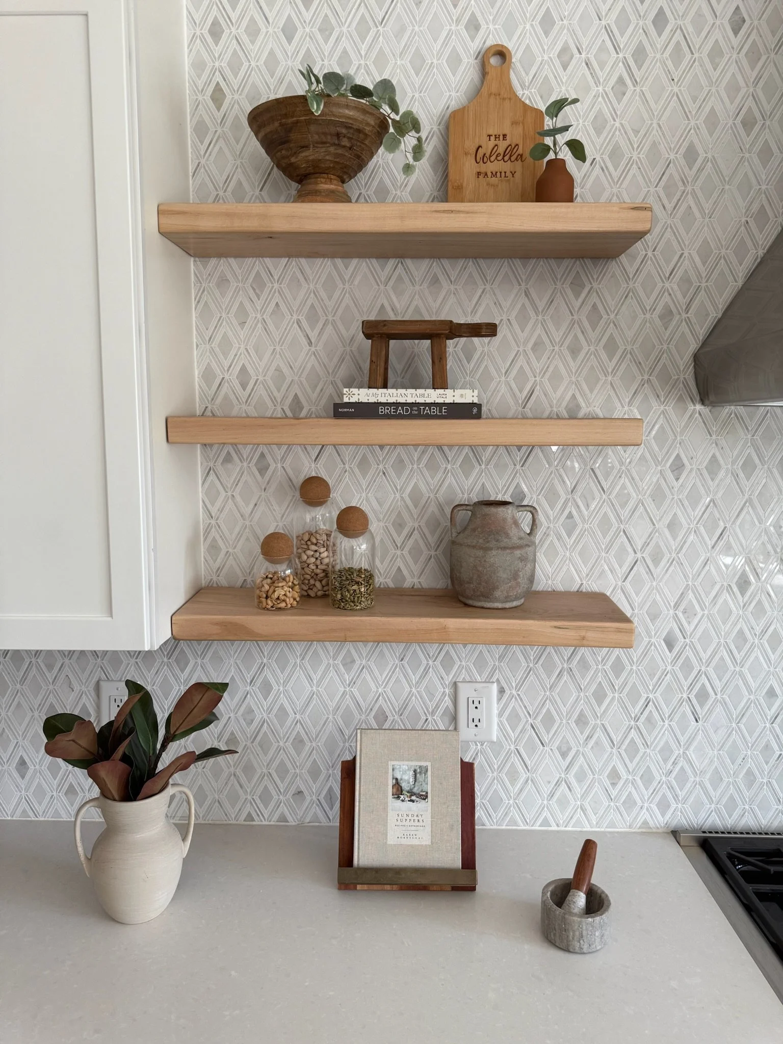 Decorative kitchen wall with three wooden floating shelves holding plants, books, jars, and pottery. A white countertop with a potted plant, a book holder with a book, and a mortar and pestle. Patterned wallpaper background.