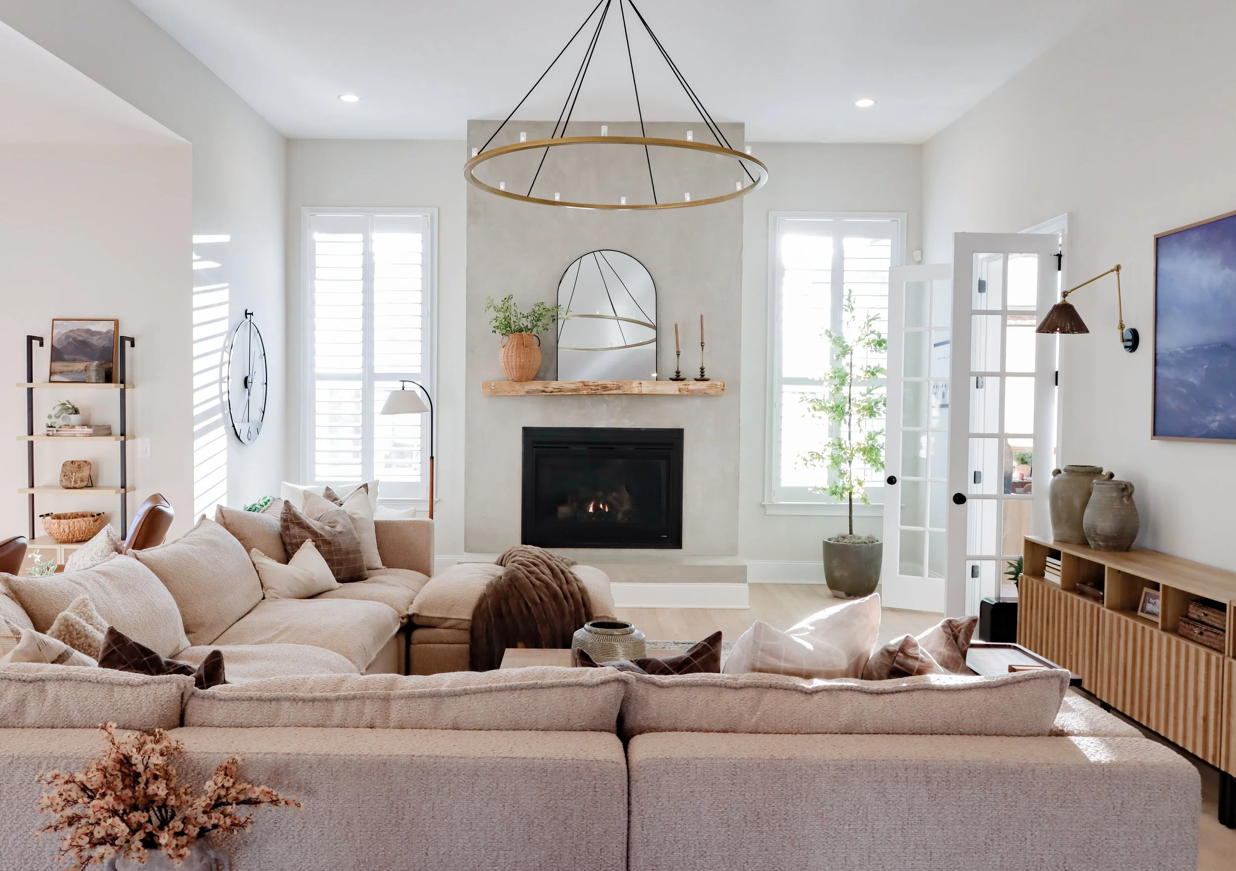 Bright living room with beige sectional sofa, fireplace, windows with shutters, wooden shelving, and modern decor.