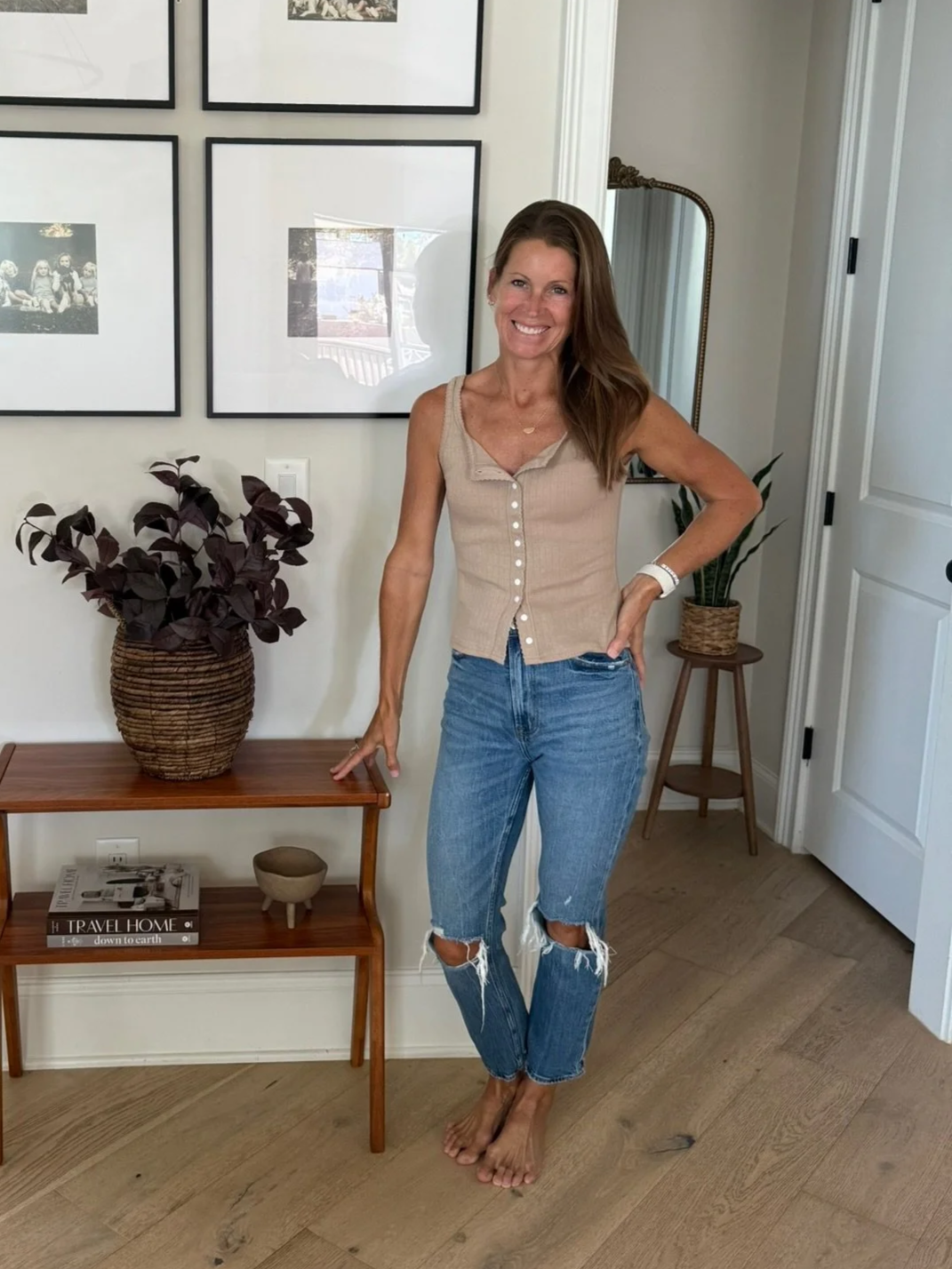 A smiling woman with long brown hair standing barefoot in a room with white walls and wooden flooring. She is wearing a beige sleeveless top and ripped blue jeans. Behind her, there is a small wooden table with a potted plant, a book, and a decorative bowl on it, and a tall plant in a wicker basket. There are four black-framed photos on the wall and a tall mirror with a gold frame beside her.