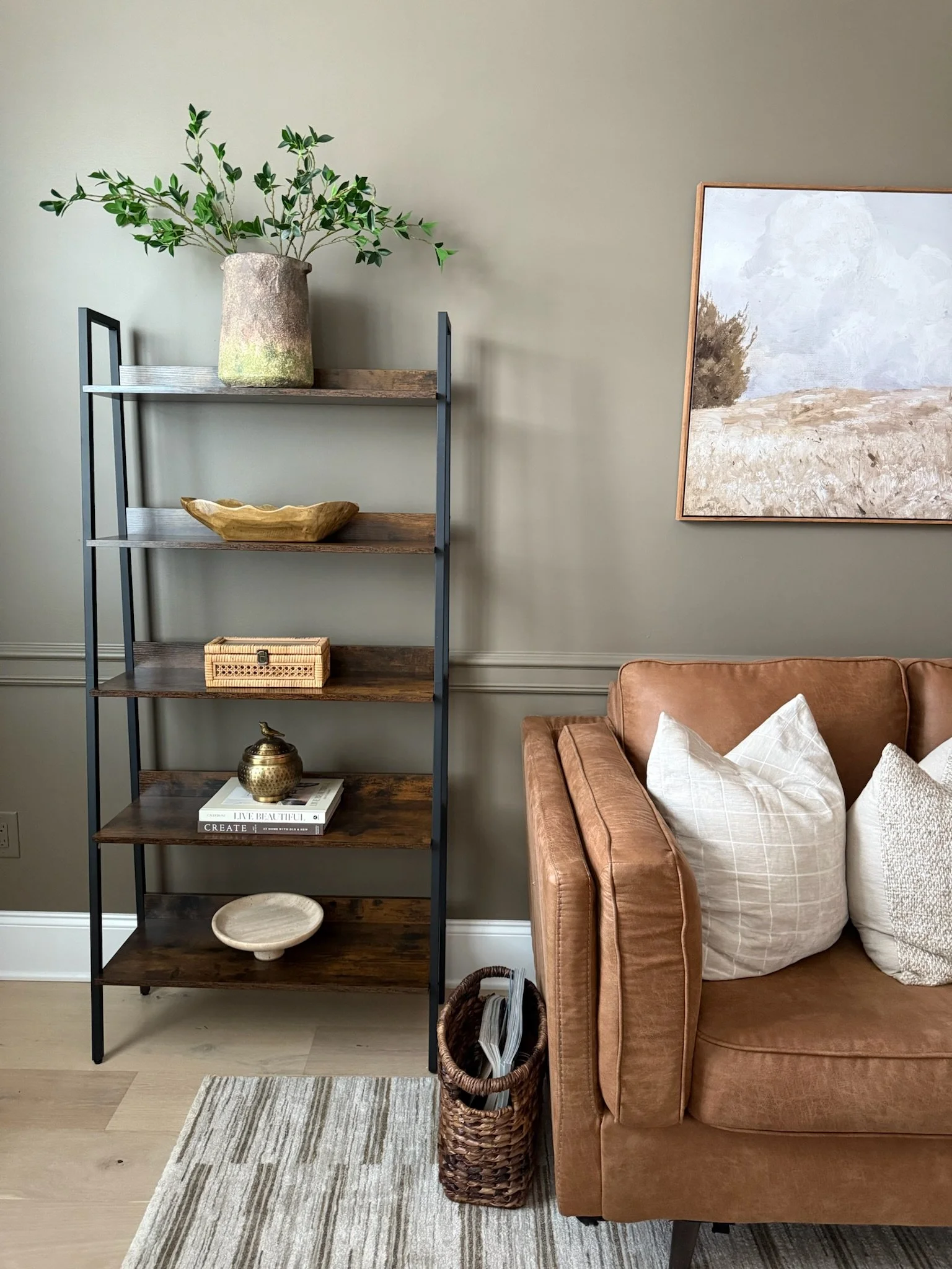 Indoor living room corner with a wooden and metal bookshelf, a tan sofa with white pillows, a basket with magazines, a small rug, and a framed landscape painting on a beige wall.