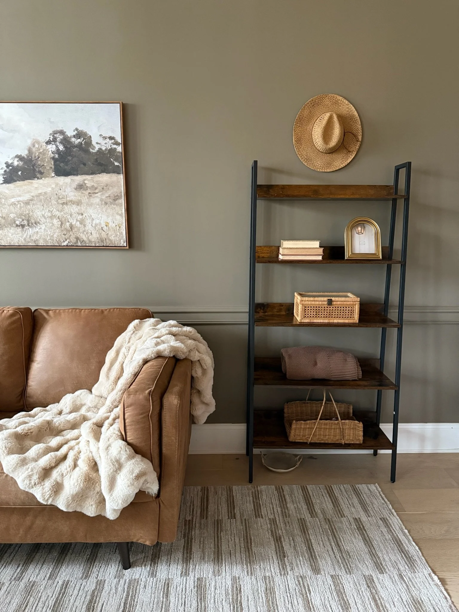 A living room corner with a brown leather sofa draped with a cream faux fur throw, next to a black metal and wood shelf with decorative items and woven baskets, beige walls, and a landscape painting hanging on the wall.