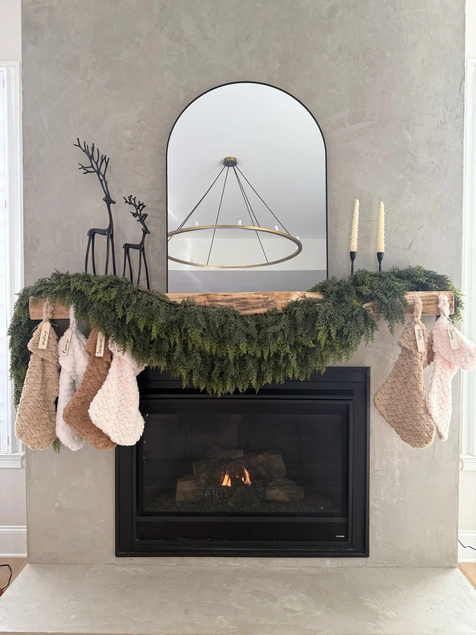 Fireplace with a wooden mantel decorated for Christmas, adorned with a green garland and hanging stockings. Above, a mirror reflects a modern circular chandelier, and on the mantle are black reindeer figurines and white candles.