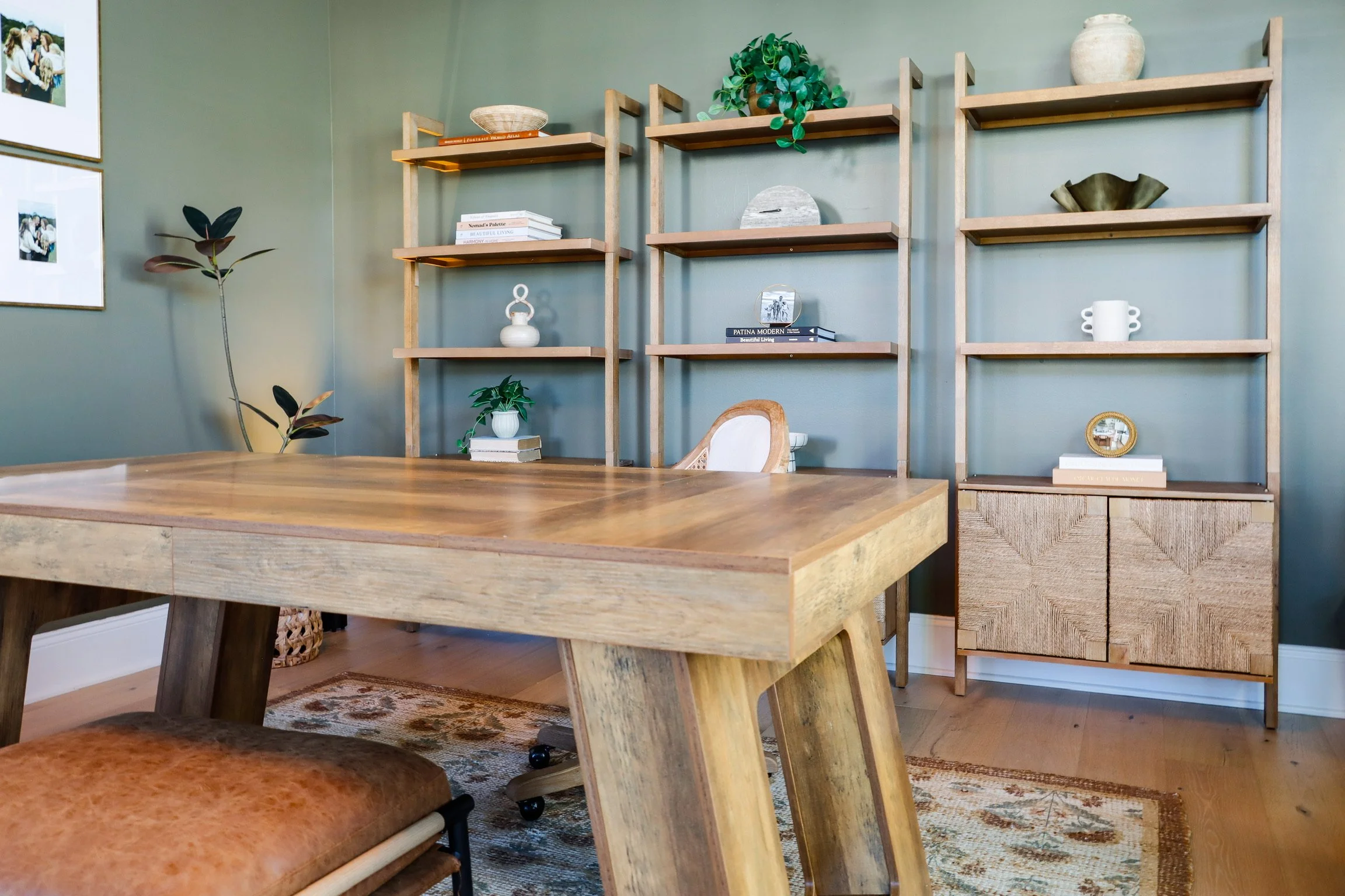 A modern home office featuring a wooden table, a rattan chair, green shelves with decorative items, potted plants, and framed photos on a green wall.