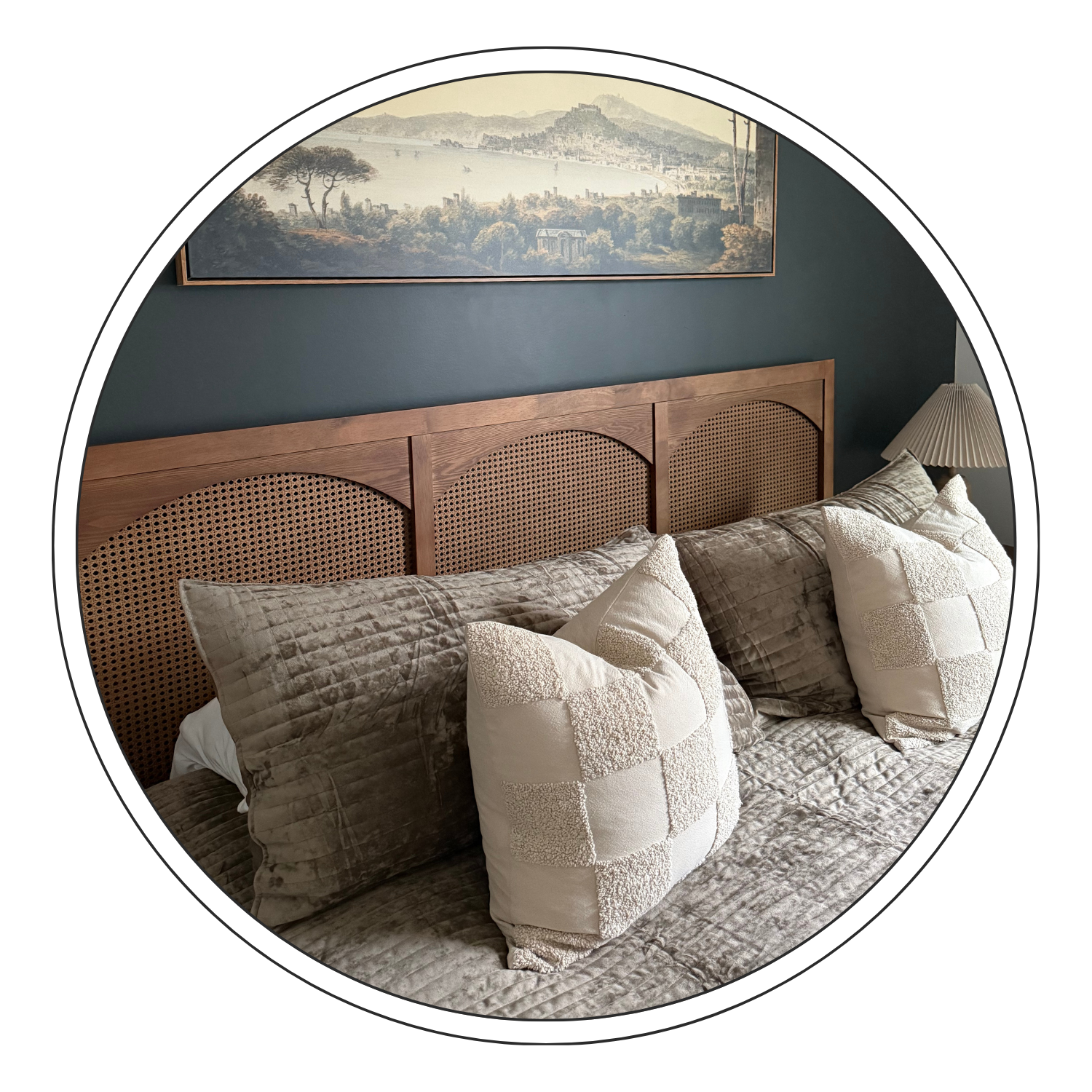 A bedroom with a wooden headboard, beige decorative pillows, dark bedside lamp, and a landscape painting on a dark green wall.
