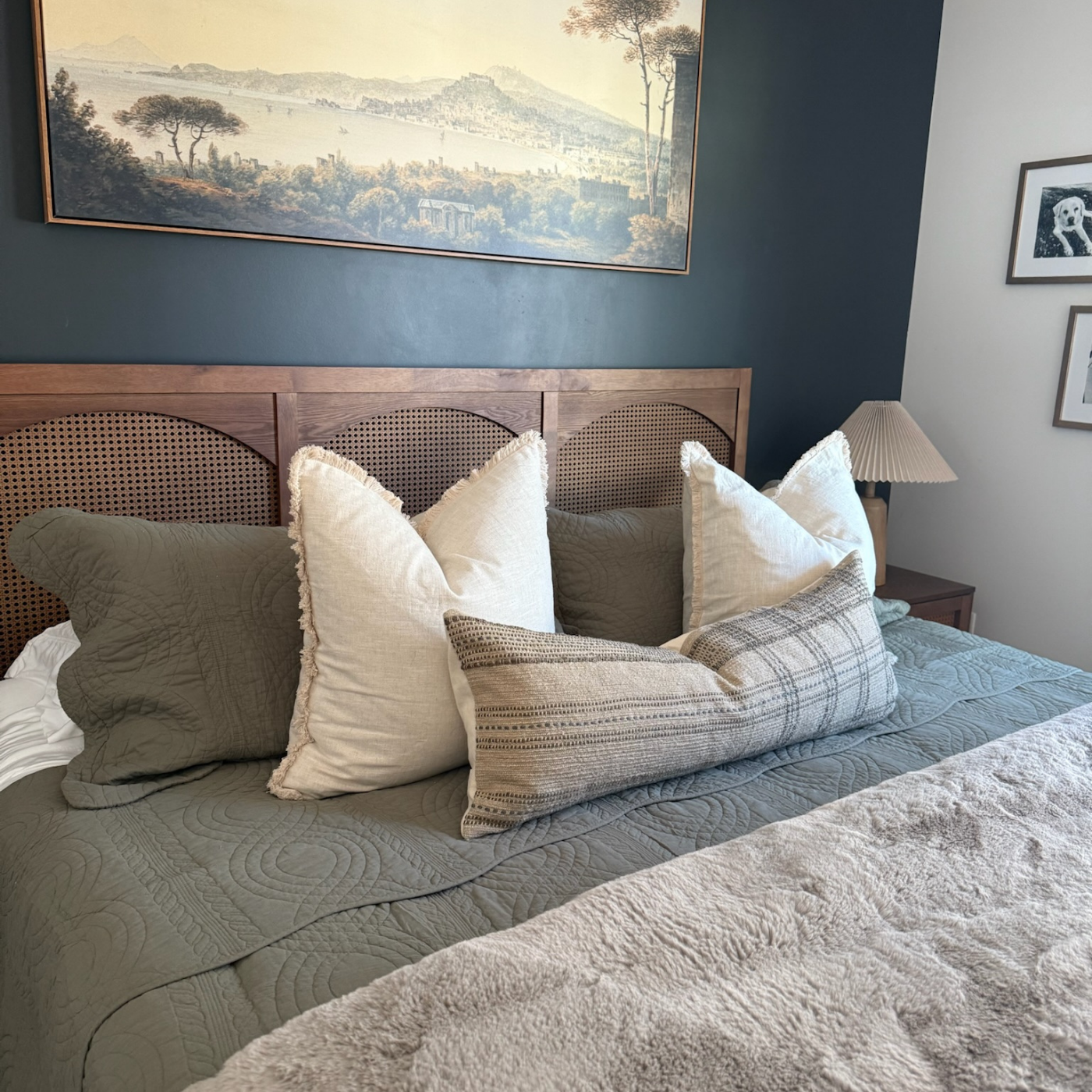 A bedroom with a wooden headboard, beige decorative pillows, dark bedside lamp, and a landscape painting on a dark green wall.